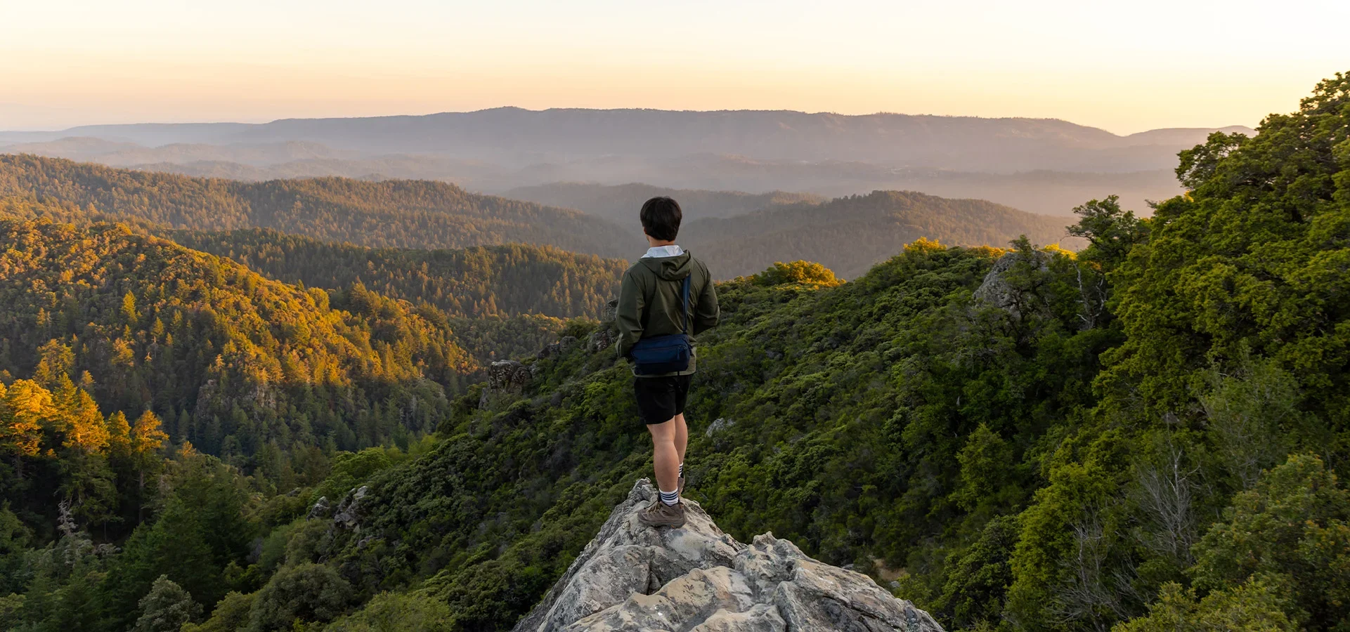 A person standing on a rocky outcrop overlooking a lush, green forested mountain landscape at sunset.