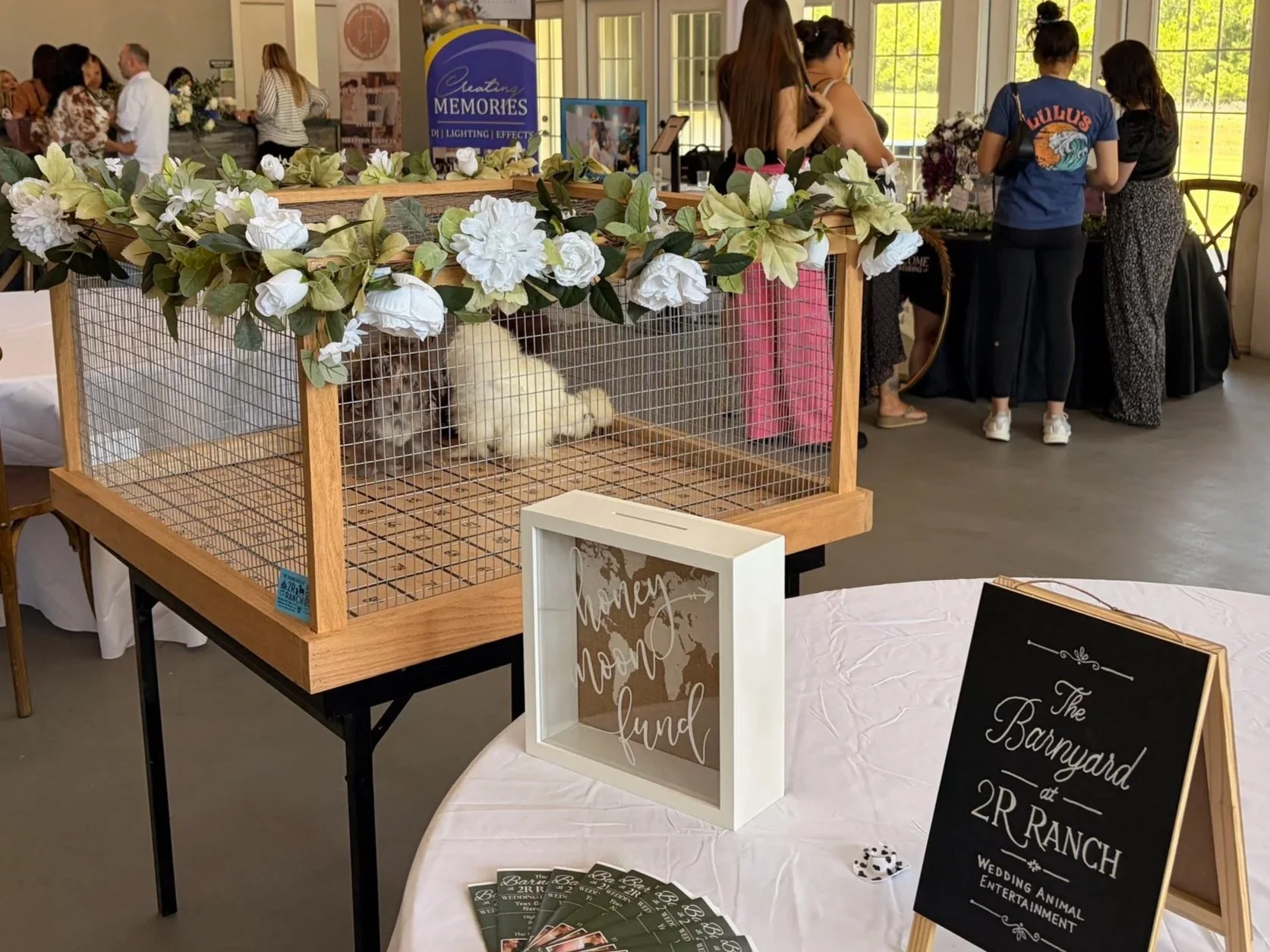 Inside an event space with a decorative enclosure with floral garland on top. In the background, people are chatting near tables and windows, and there is a sign for the Barnyard at 2R Ranch, indicating a wedding animal entertainment event.