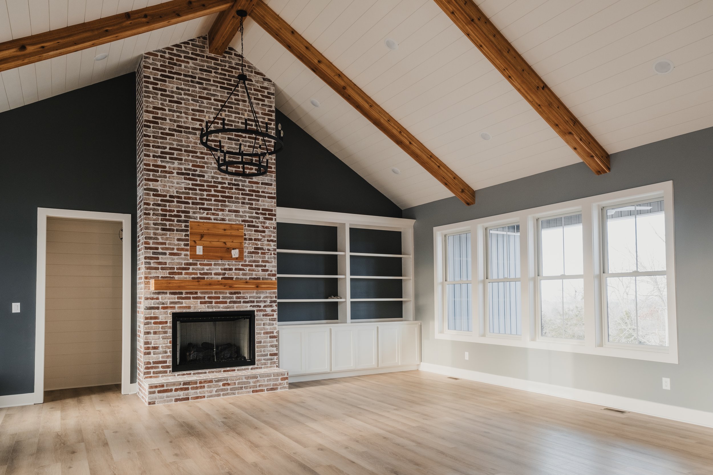 Empty living room with brick fireplace, built-in shelving, large windows, a vaulted ceiling with exposed wooden beams, and light wood flooring.
