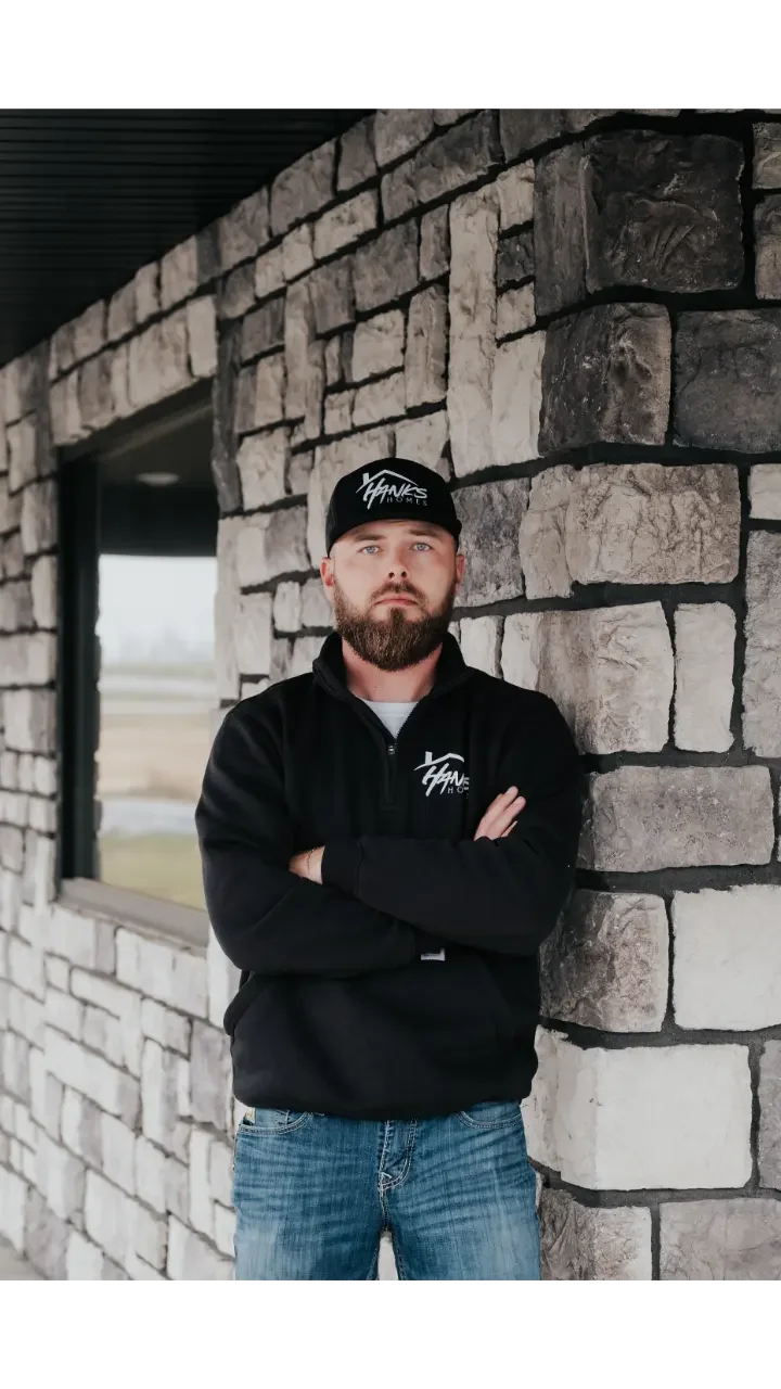 A man with a beard wearing a black cap and black jacket with white logo, standing with arms crossed against a stone wall near a window in an outdoor setting.