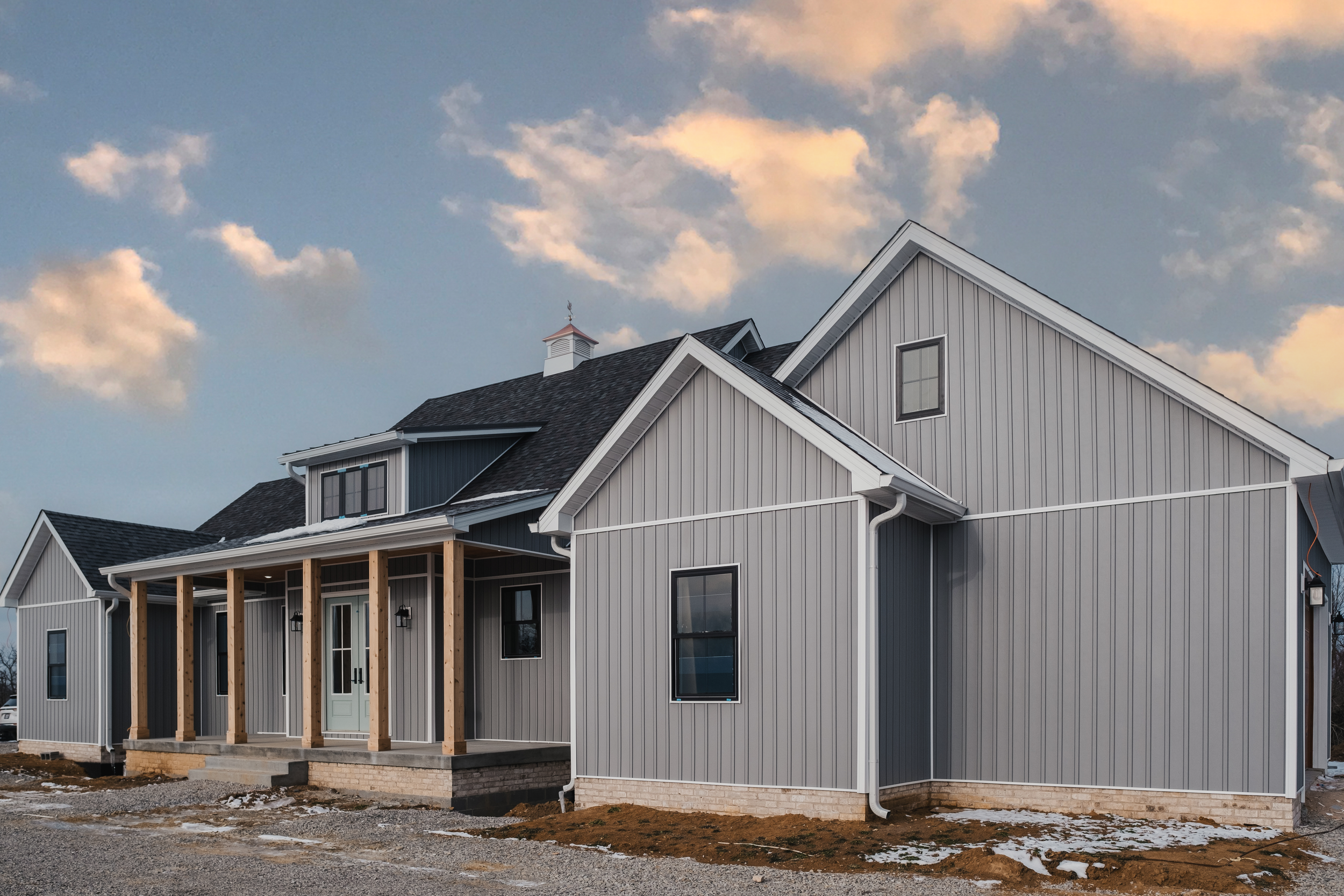 Newly built modern house with gray vertical siding and wooden porch columns, under a partly cloudy sky at sunset.