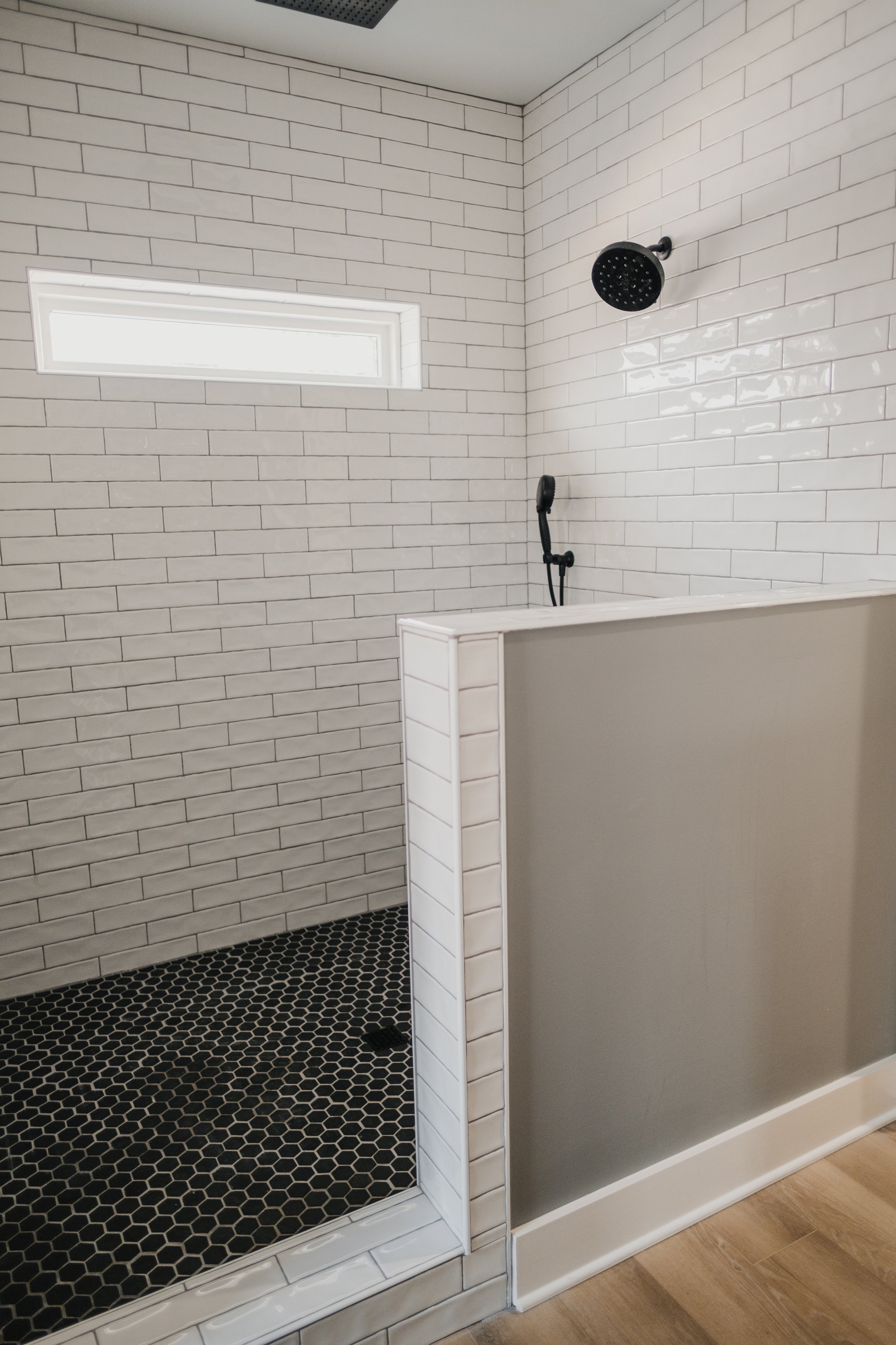 Modern shower with white subway tiles, a black rain showerhead, a handheld shower, a small horizontal window, black hexagonal floor tiles, and partial wall divider.