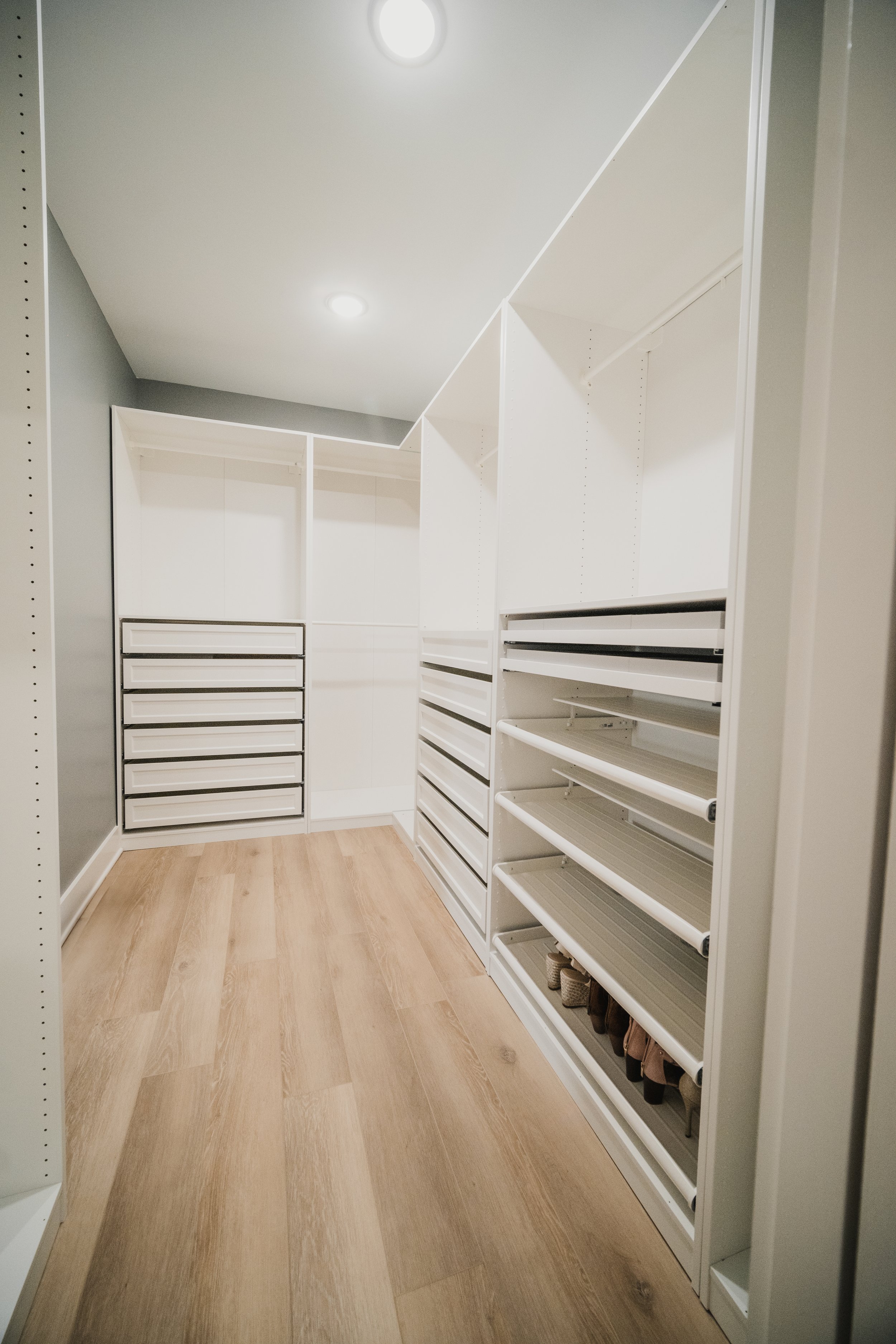 Empty walk-in closet with white shelving units and drawers, wood flooring, and white ceiling lights.