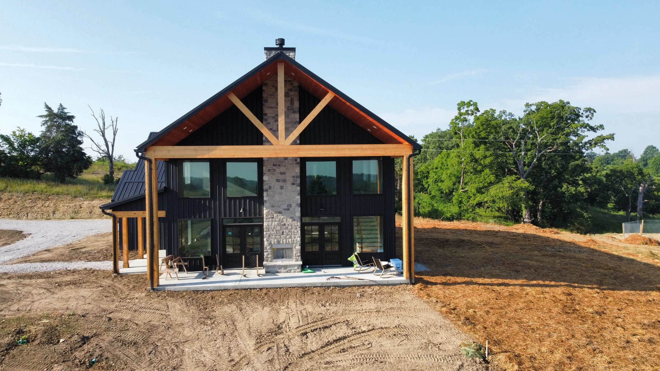 Modern two-story house under construction with black exterior, wooden accents, and large windows, surrounded by a cleared yard and green trees in the background.