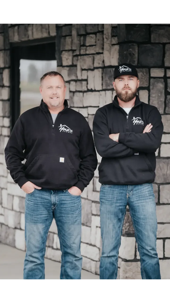 Two men standing in front of a stone wall, wearing black jackets with a logo that says 'Hanks Homes', one with hands in pocket and the other with crossed arms, both looking at the camera.