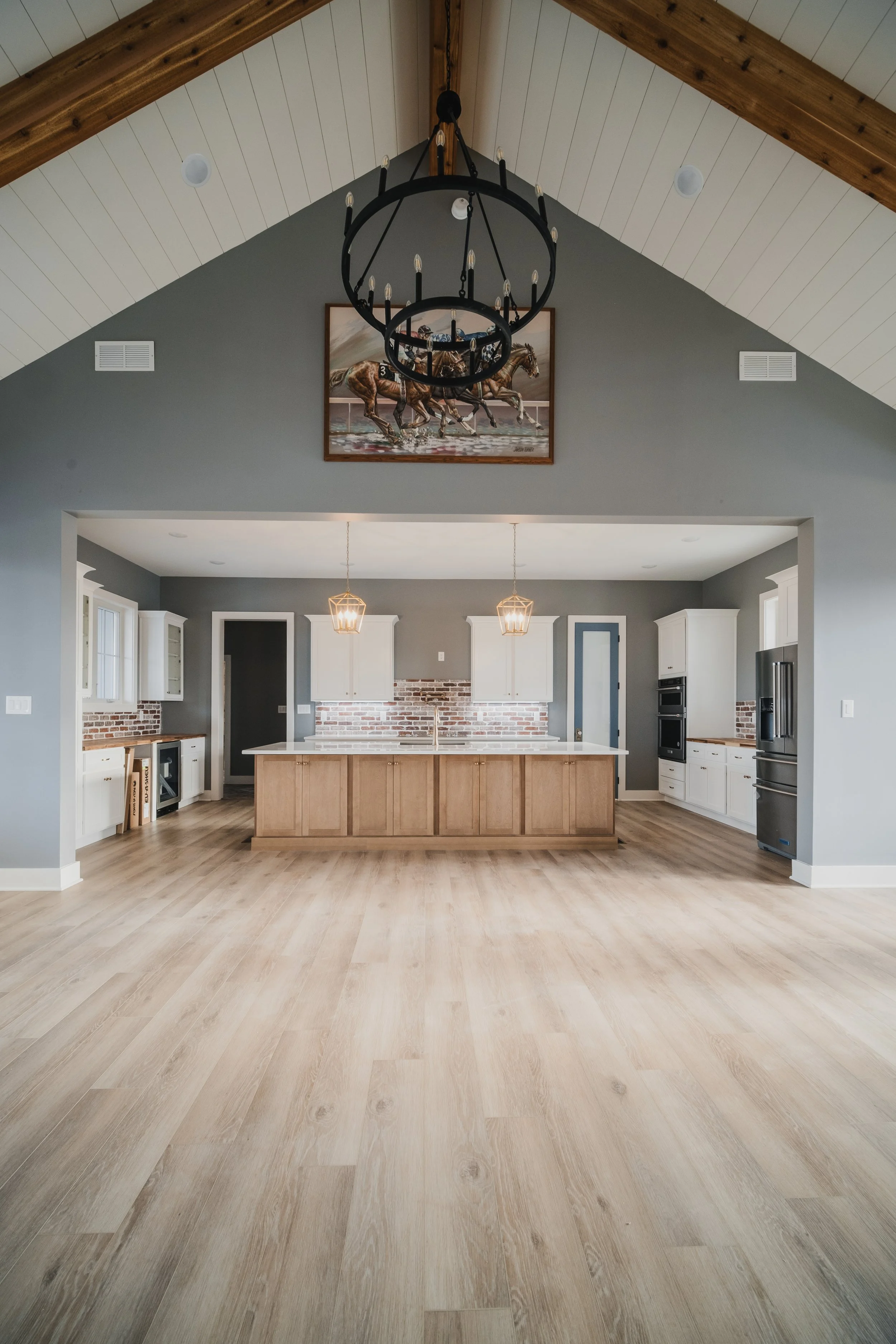 Spacious kitchen with light wooden floors, white cabinets, brick backsplash, and a large island in the center, under a vaulted ceiling with wood paneling and a black chandelier.