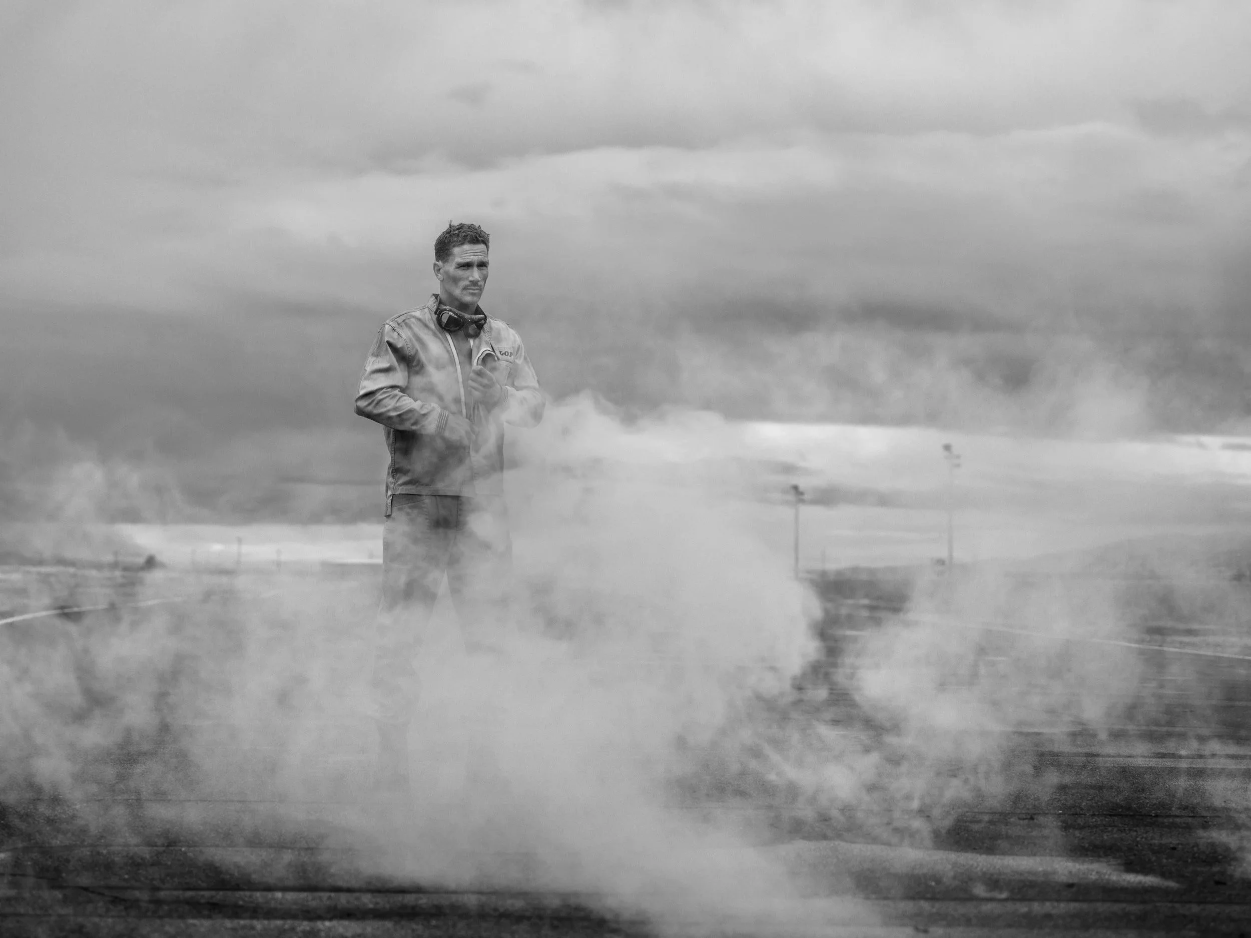 A man standing on an airstrip surrounded by smoke or vapor, wearing a jumpsuit with a headset around his neck, looking into the distance with a serious expression.