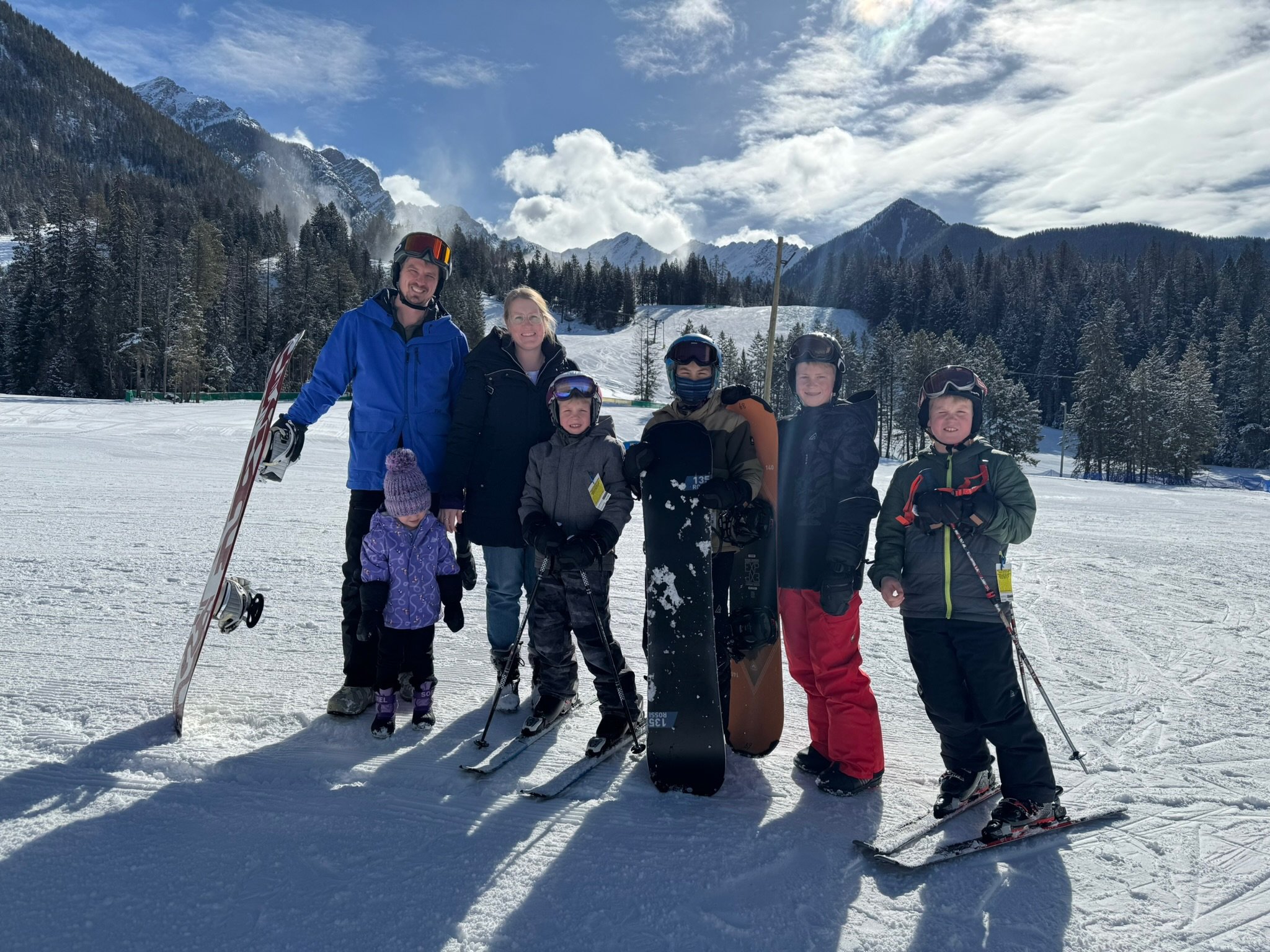 A group of seven people, including children and adults, in winter clothing on a snowy mountain with ski equipment, trees, and mountains in the background.