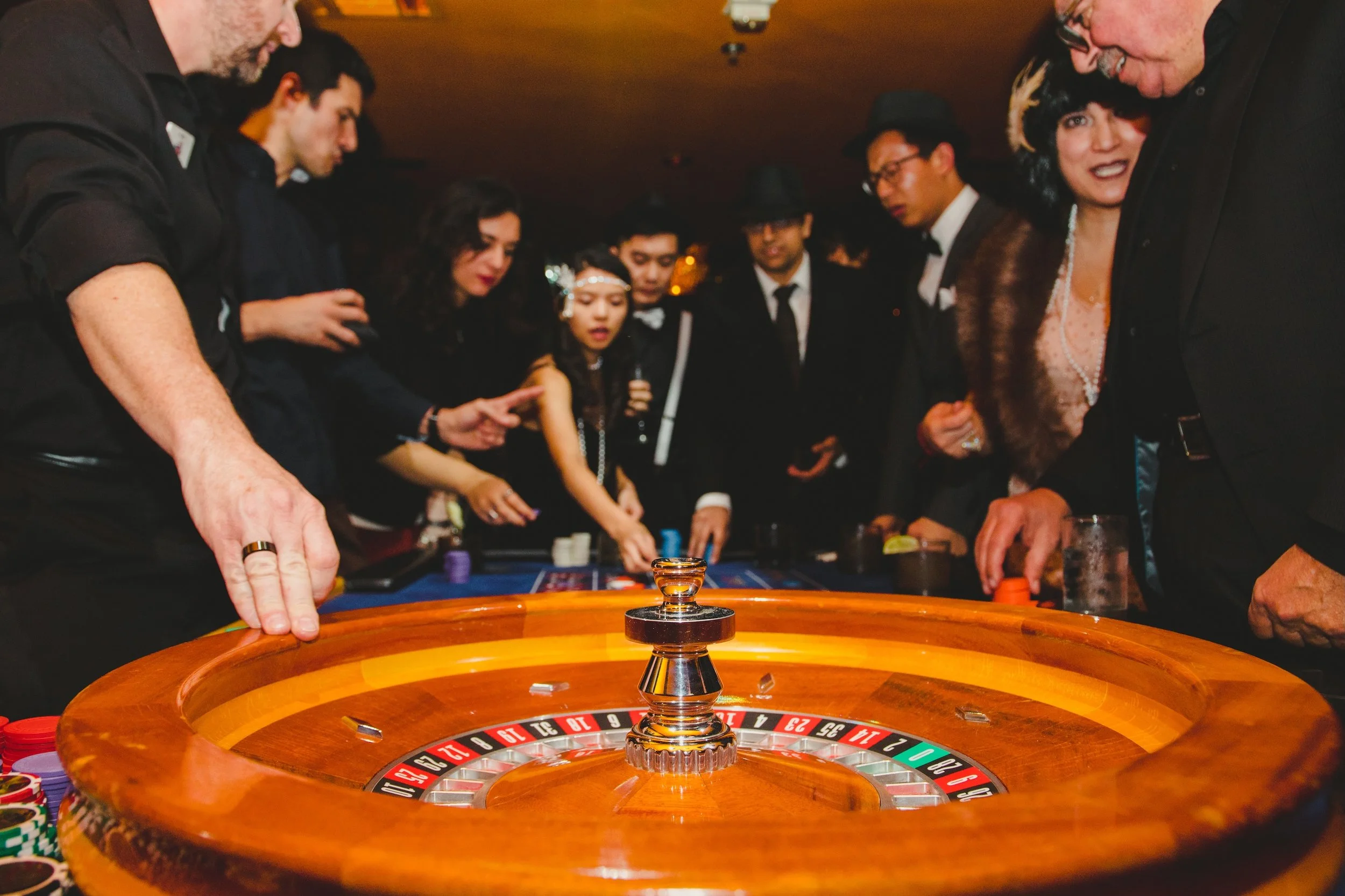 People gathered around a roulette table playing casino game at a party or casino, with chips and drinks on the table.
