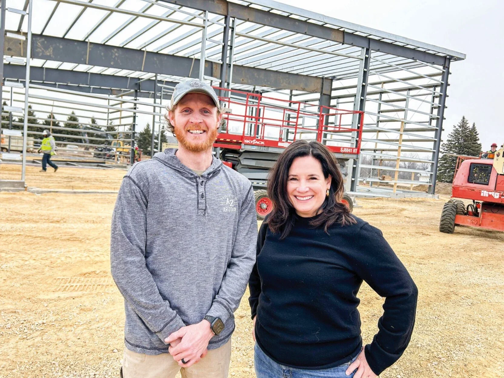 Two smiling people standing outdoors at a construction site with metal framework and construction equipment.