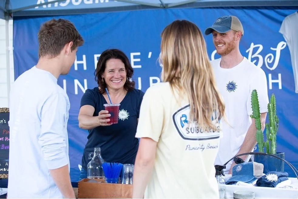 Four people at a booth with a blue banner, two women and two men, one woman holding a red drink, smiling, with cactus plants on the side.