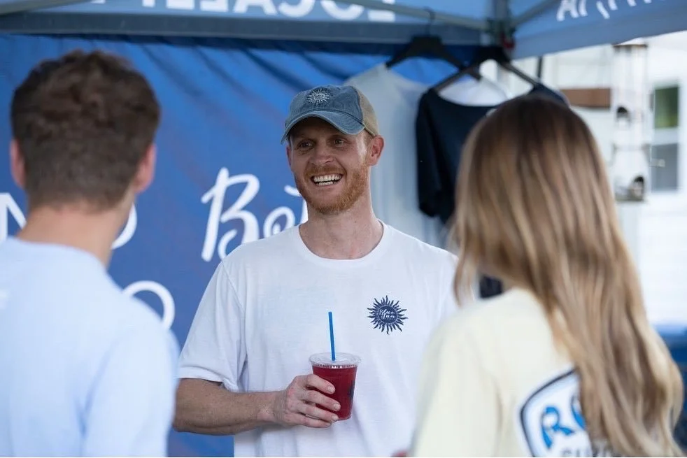 A man with a beard and gray cap smiling and holding a red drink with a blue straw, talking to two people at an outdoor event or festival.