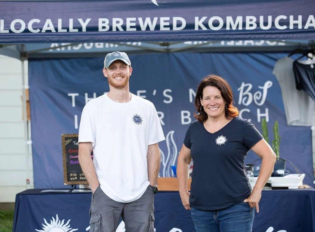 Two people, a man and a woman, standing and smiling at an outdoor booth that sells locally brewed kombucha. The booth has a navy blue canopy with white text and a matching backdrop featuring a sun logo.