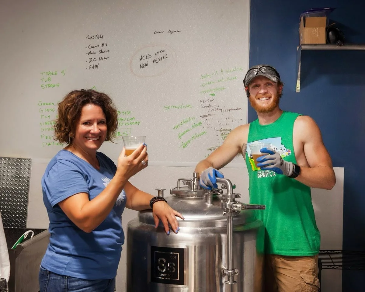 A woman and a man are smiling and holding glasses of beer while standing next to a stainless steel brewing vessel in a brewery or brewpub. The woman has curly brown hair and is wearing a blue t-shirt, and the man has a beard, is wearing a green tank top, gloves, and a cap. In the background, there is a whiteboard with notes and a shelf with some items.