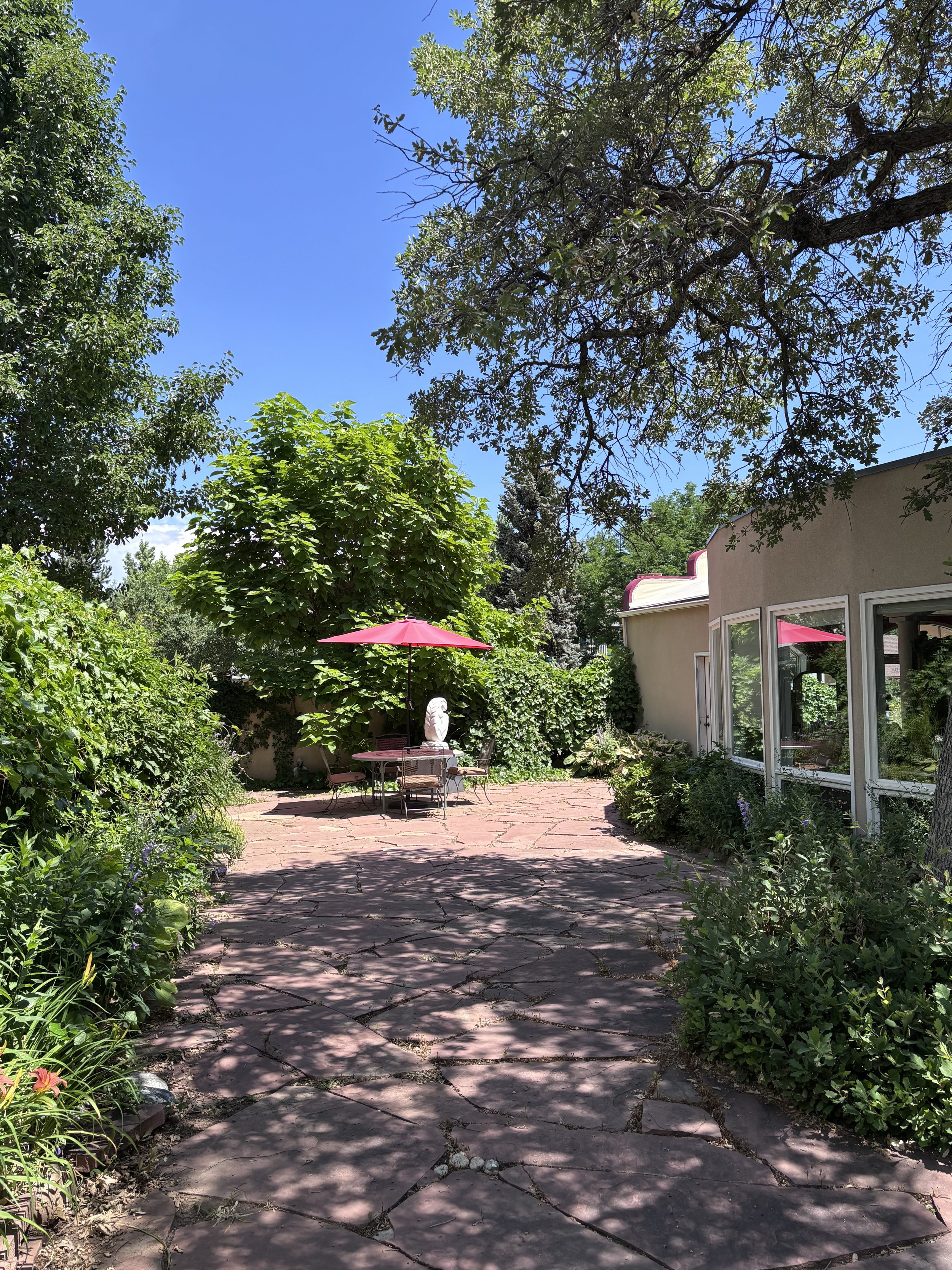 A sunny backyard patio scene with a stone pathway leading to a patio table and chairs, shaded by a pink umbrella. There are lush green trees and bushes, with a house visible on the right side, and a clear blue sky overhead.