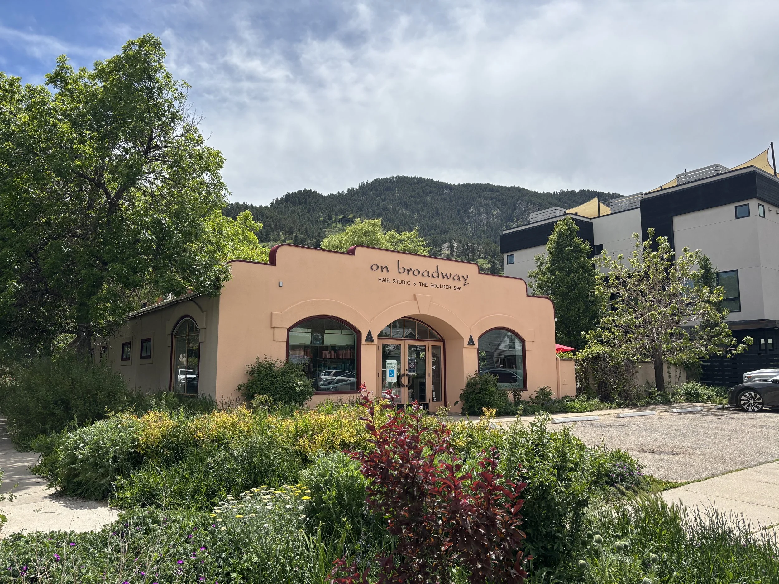 A pink building with a sign reading 'on broadway, hair studio & the boulder spa,' surrounded by green plants and trees, with mountains and a partly cloudy sky in the background.