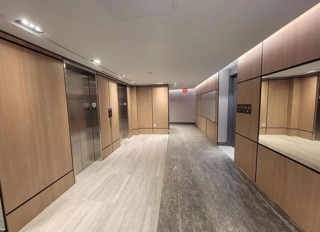 Empty corridor with two elevators on the left, wood-paneled walls, and a mirrored wall reflection on the right, in a modern office building.