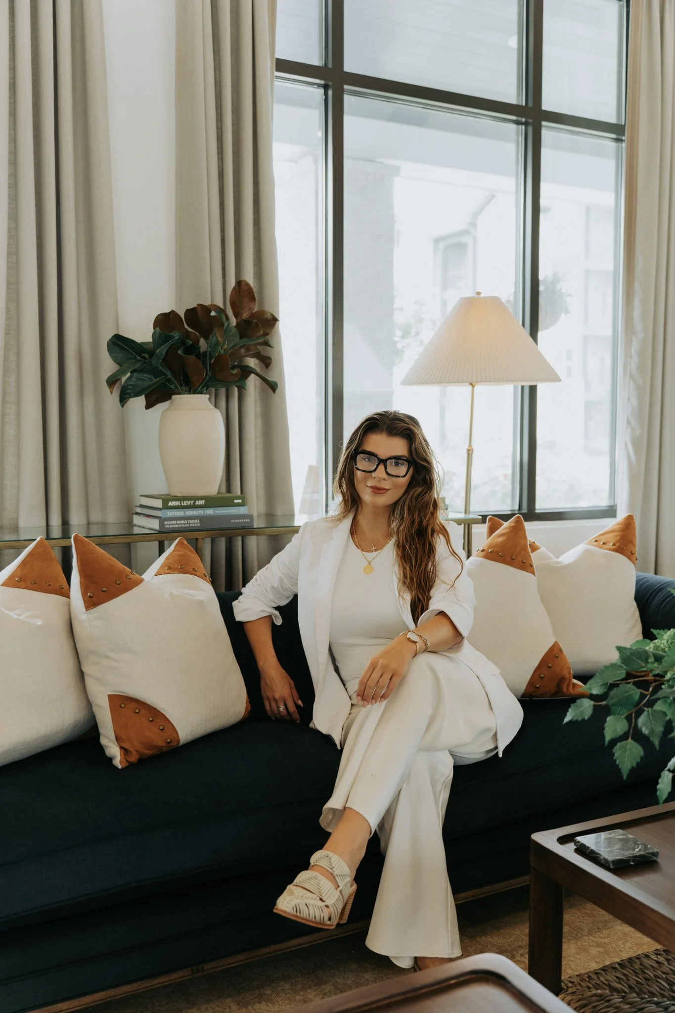 A woman with wavy brown hair and glasses sitting on a black couch decorated with white pillows with brown accents, in a modern living room with large curtains and a window showing a building outside, a white vase with large green and brown leaves, a bedside table with a lamp, and a stack of books.
