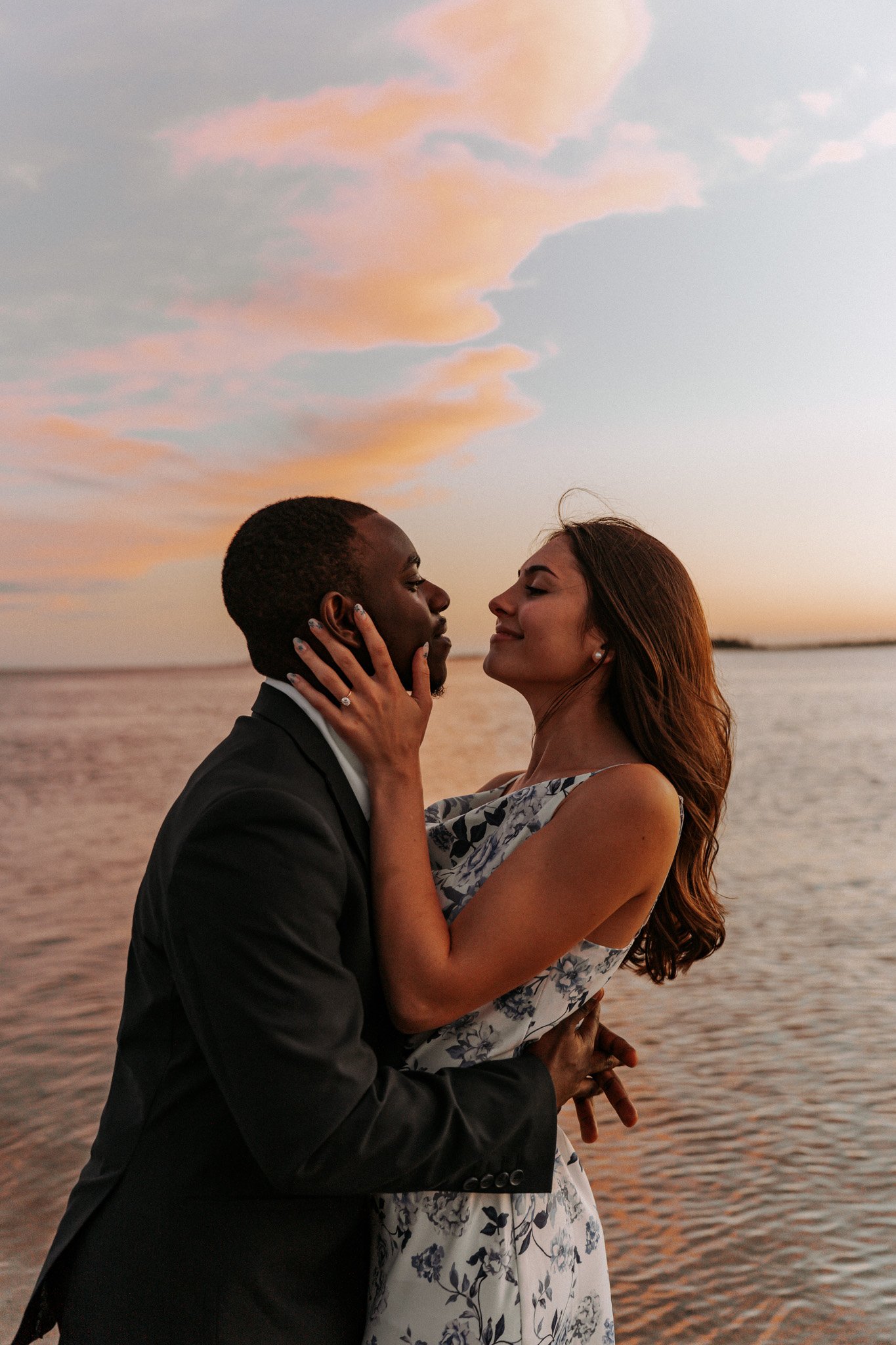 A couple standing close together by a body of water during sunset, with pastel-colored clouds in the sky. The man is wearing a dark suit, and the woman is in a floral dress. They are gazing into each other's eyes, with the woman holding the man's face and the man holding the woman's waist.