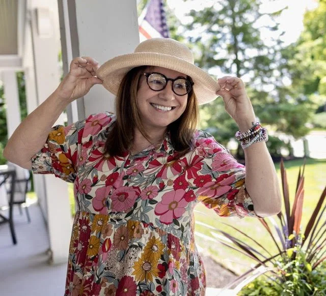 Georganne Bender smiling and wearing a wide-brimmed straw hat, glasses, a floral dress, and bracelets, standing outdoors on a porch with greenery in the background.
