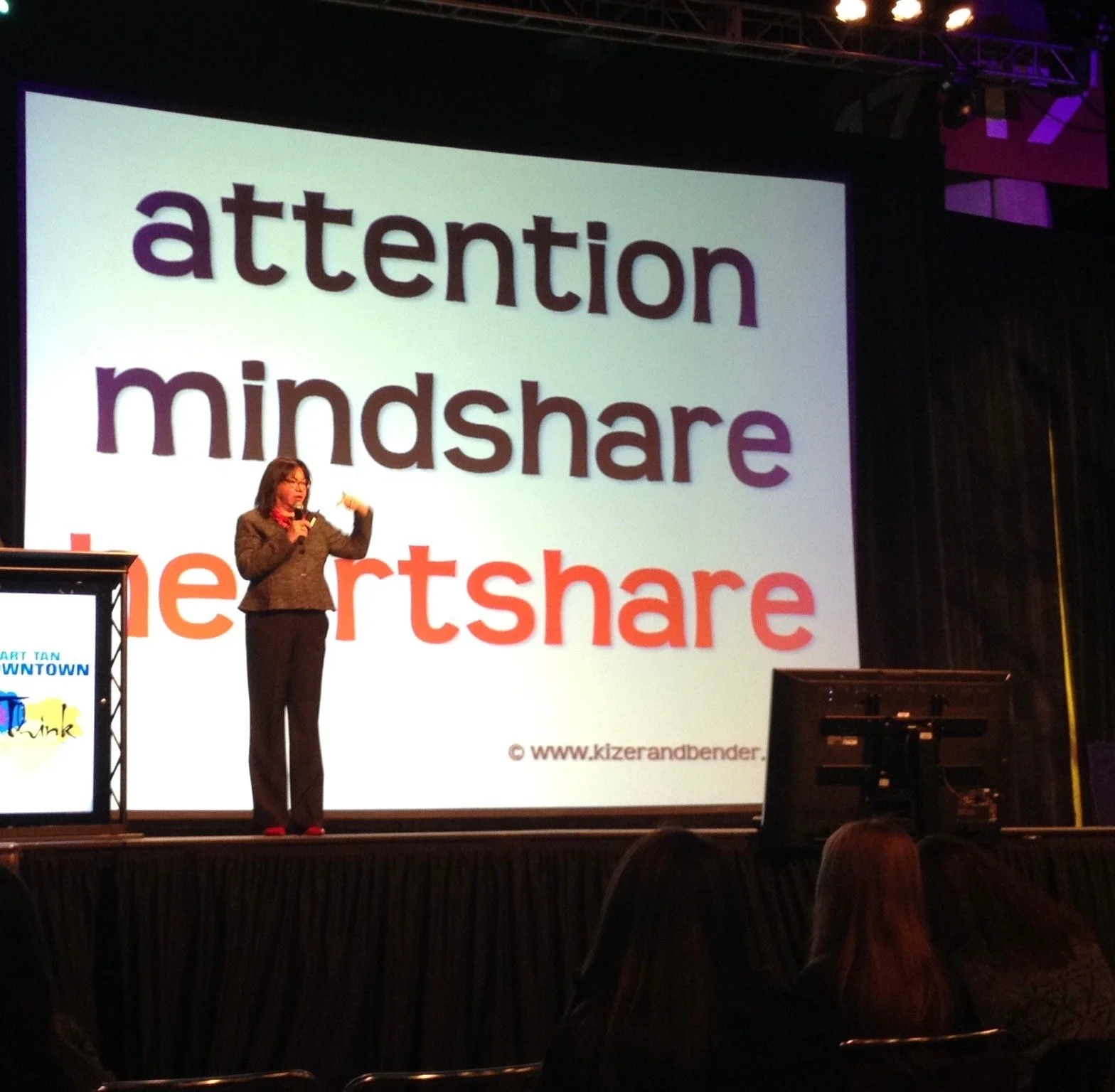 A woman in business attire is giving a presentation on stage with a large screen behind her displaying the words 'attention', 'mindshare', and 'heartshare' in large colorful letters. The stage is set with lecterns and audience members are seated in front.