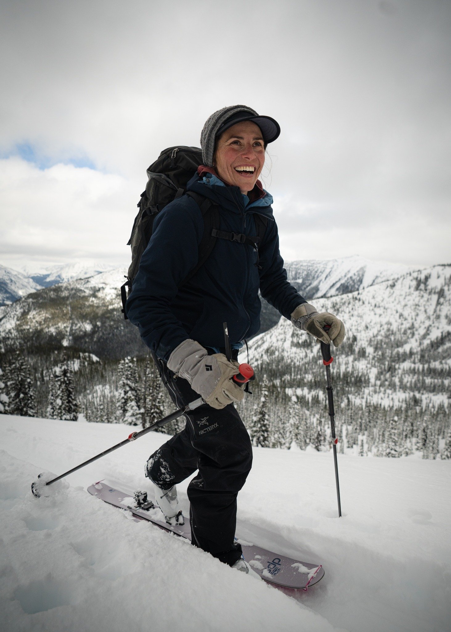 A woman snowboarding on a snowy mountain with trees and mountains in the background, smiling and dressed in winter gear.