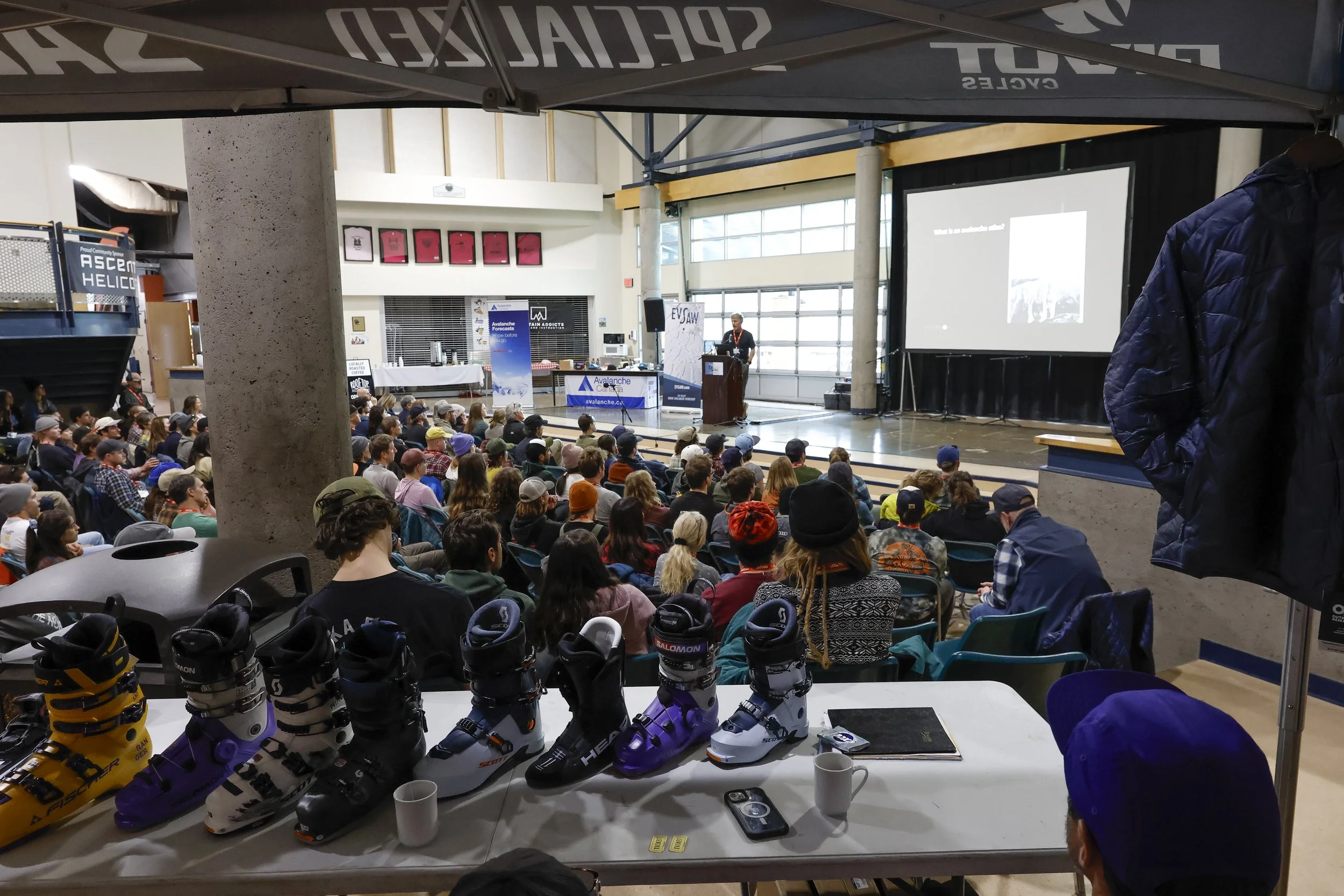 Indoor conference or seminar with a large audience seated facing a stage. The stage has a speaker at a podium, a large projection screen, and various banners. In the foreground, a table displays multiple pairs of ski boots, with some coffee cups and a cellphone placed on the table.