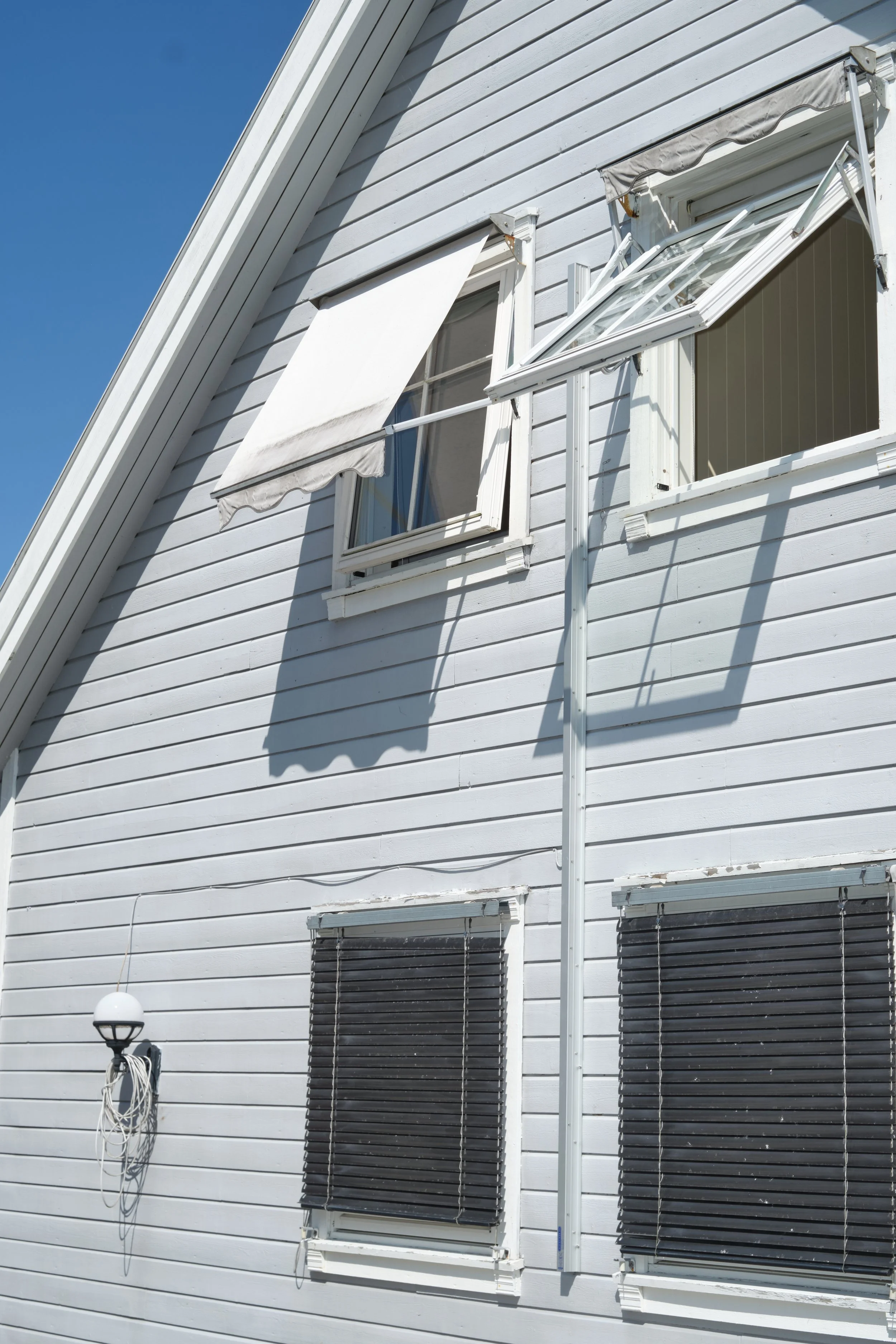 Second story of a white house with three windows, two with closed black blinds and one with an open flower box and an awning, with a rain gutter and downspout, and a streetlight with wires tied to it, under a clear blue sky.