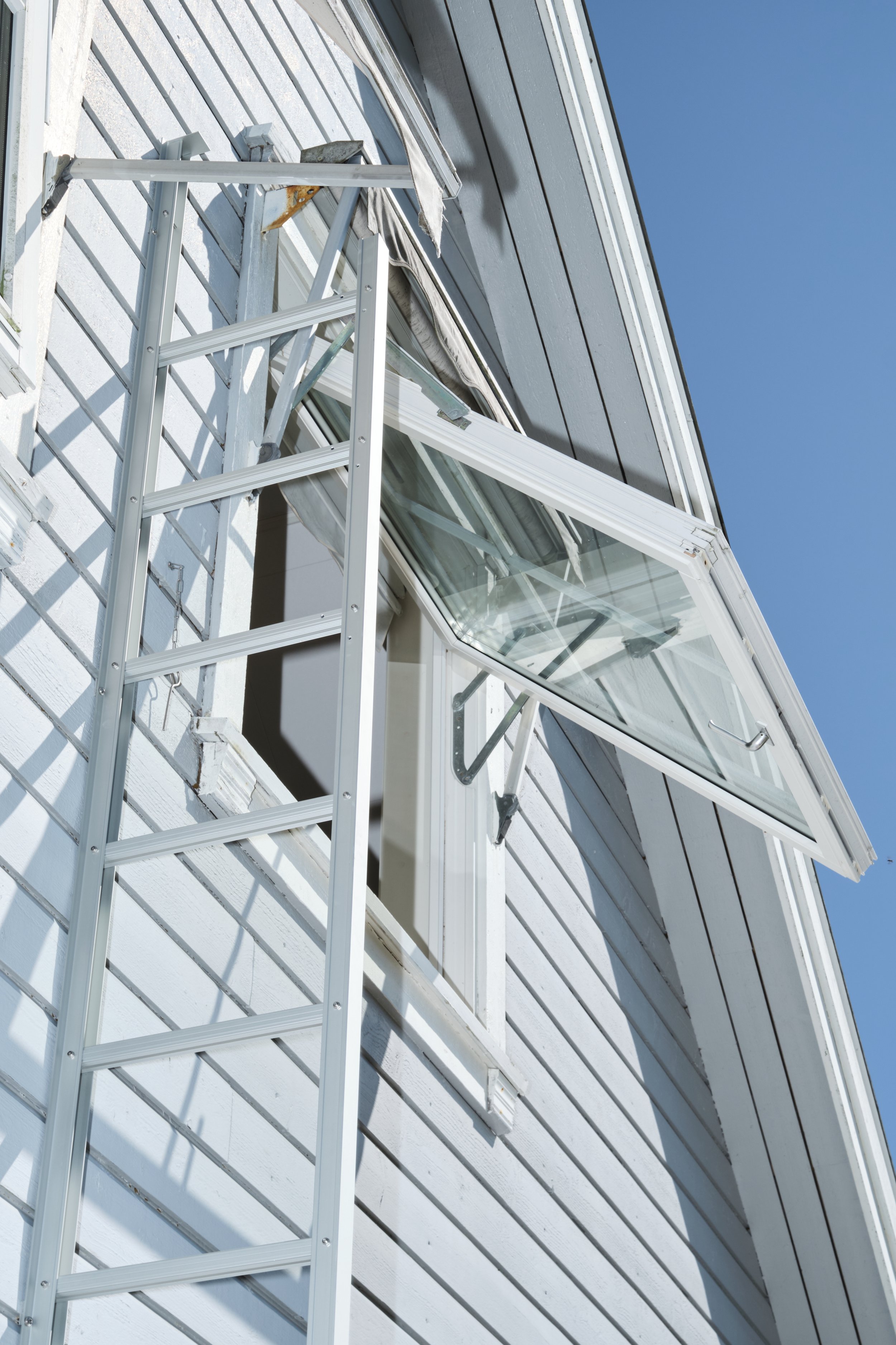 Photo of a house with a window open and a silver ladder propped against the white wooden siding, with the ladder extending to the open window on a clear day.