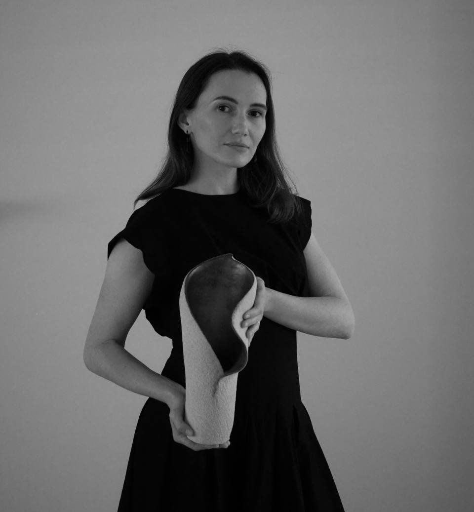 A woman in a black dress holding a large, abstract ceramic sculpture made in biophilic design, standing against a plain wall.