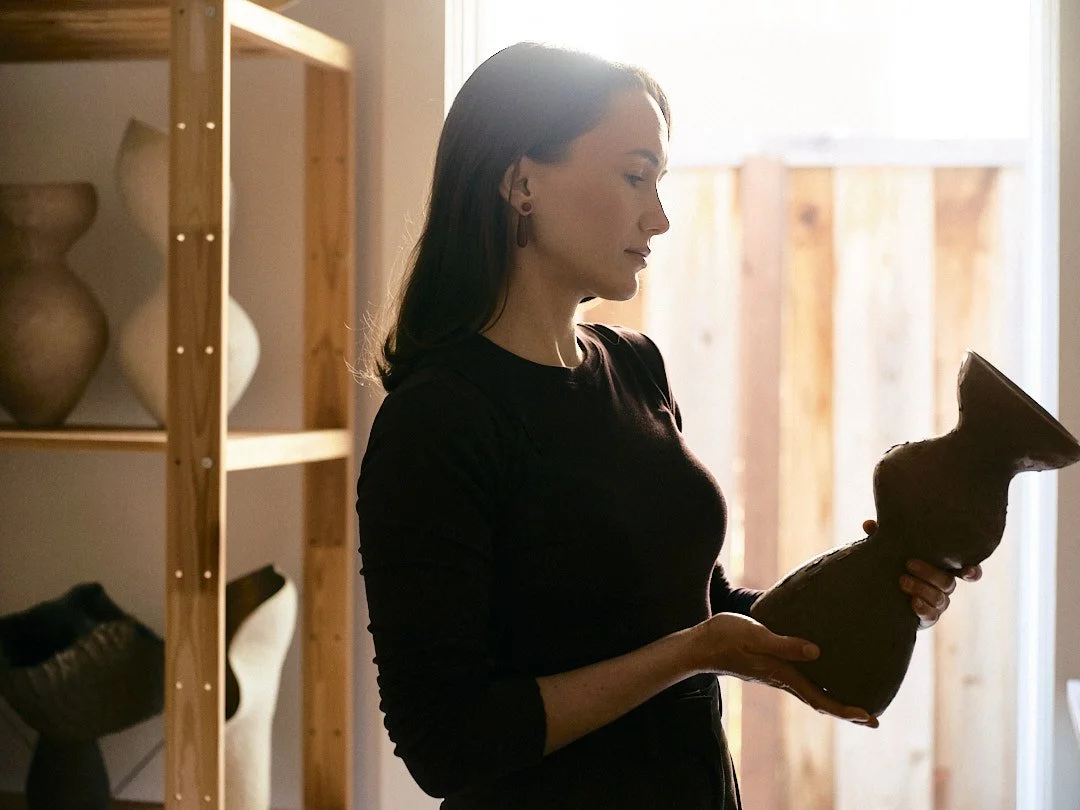 A woman in a black shirt holding a large, textured, brown ceramic vase or sculpture while standing indoors near a window.