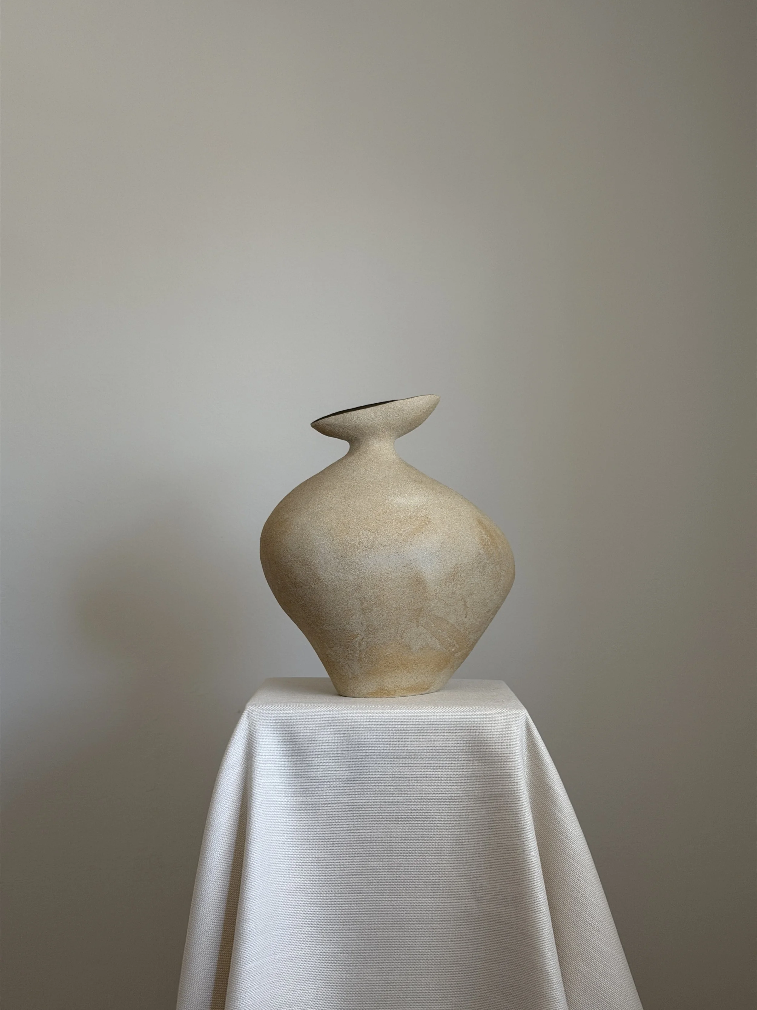 Hand-built cream stoneware vase with a rounded body and flared, offset mouth on a draped plinth