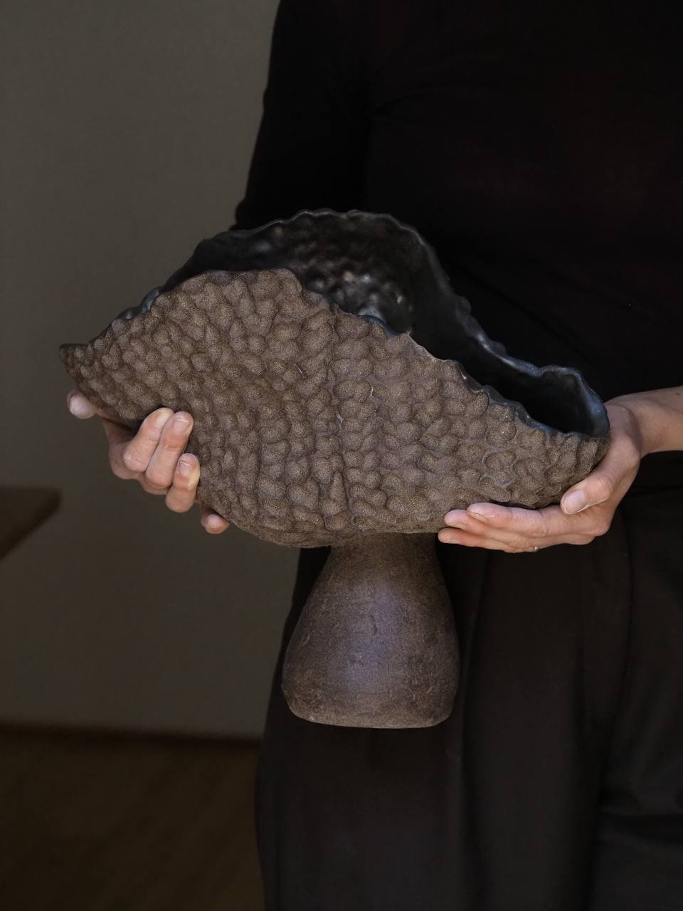 Person holding a large organic shaped vase made in biophilic design with a rounded edge, and made from textured brown clay