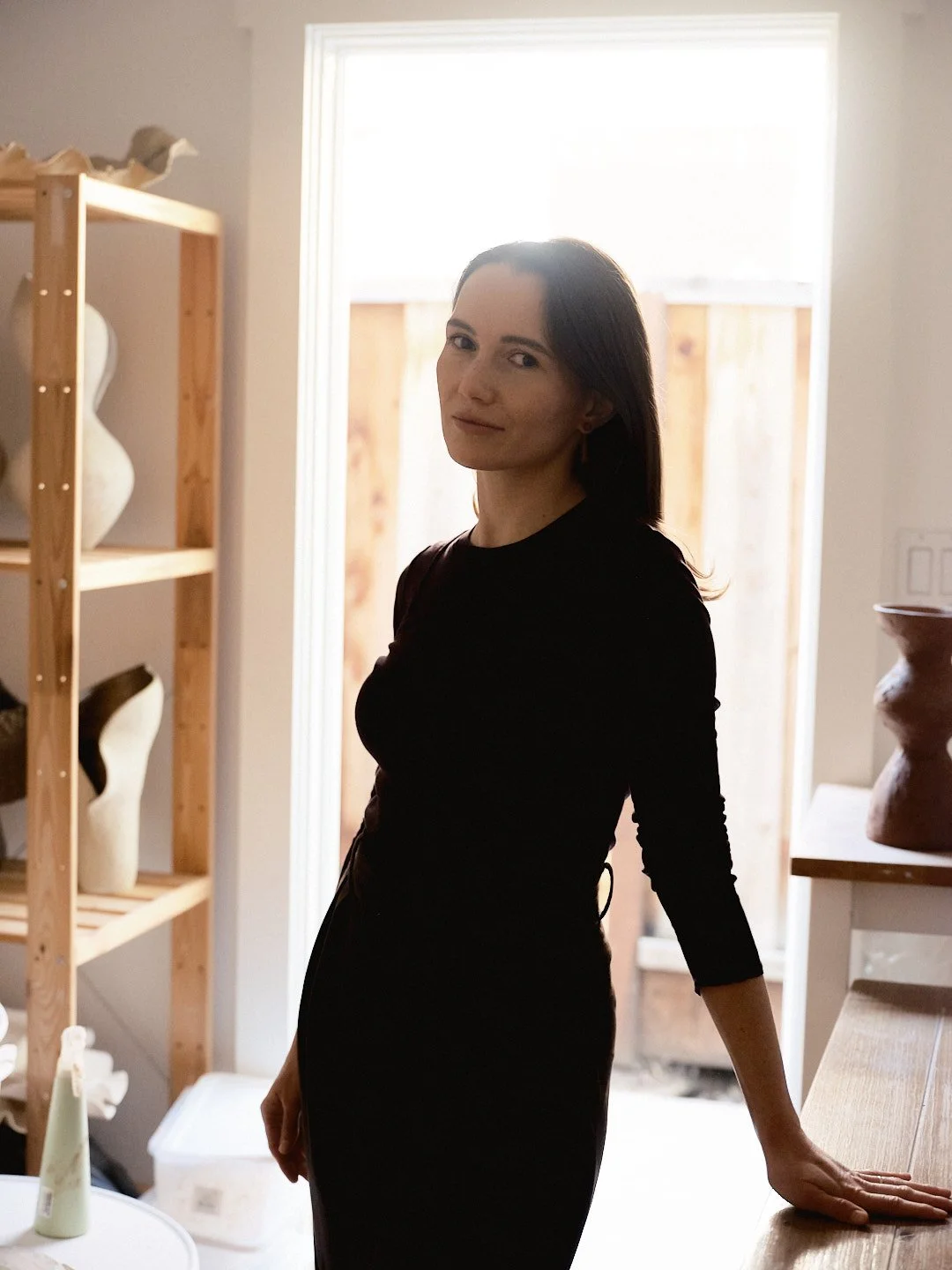A woman with shoulder-length dark hair and fair skin standing indoors with natural light, wearing a black long-sleeve dress, in a room with wooden shelves holding white and beige vases, and a window with an outdoor view of a wooden fence.