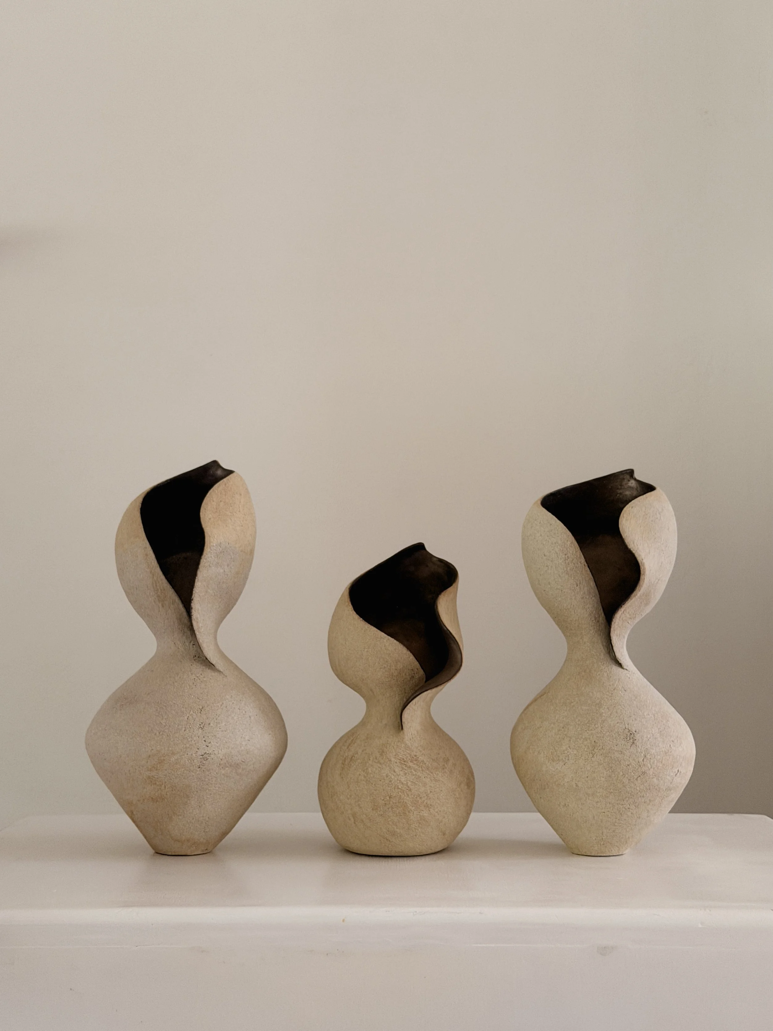 Three cream sculptural ceramic vases in graduated sizes, with dark interiors and curved wave-like openings.