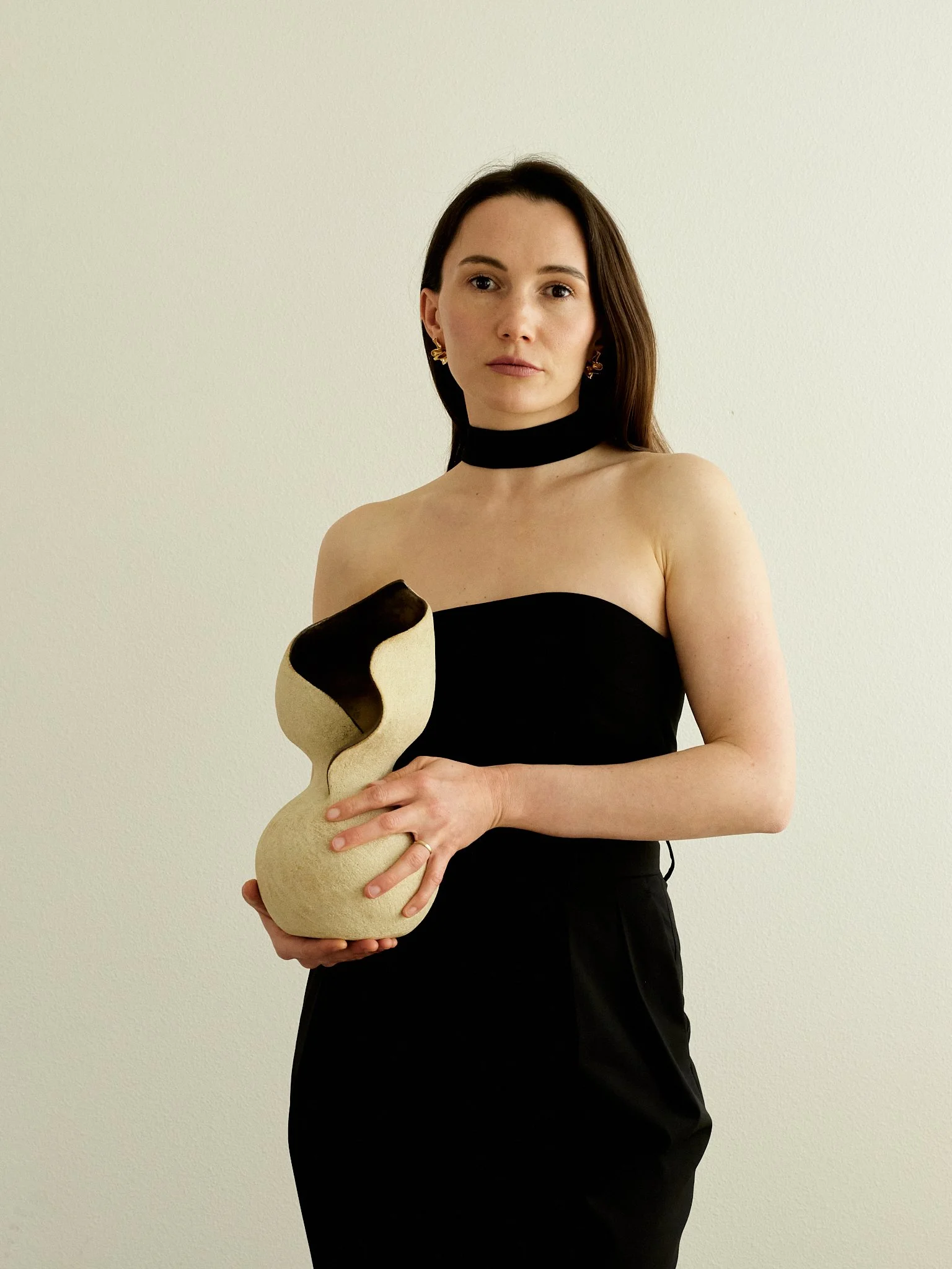 Anastasia Babenko holding a sand-colored hand-built sculptural ceramic vessel with a wave-cut rim and dark glazed interior