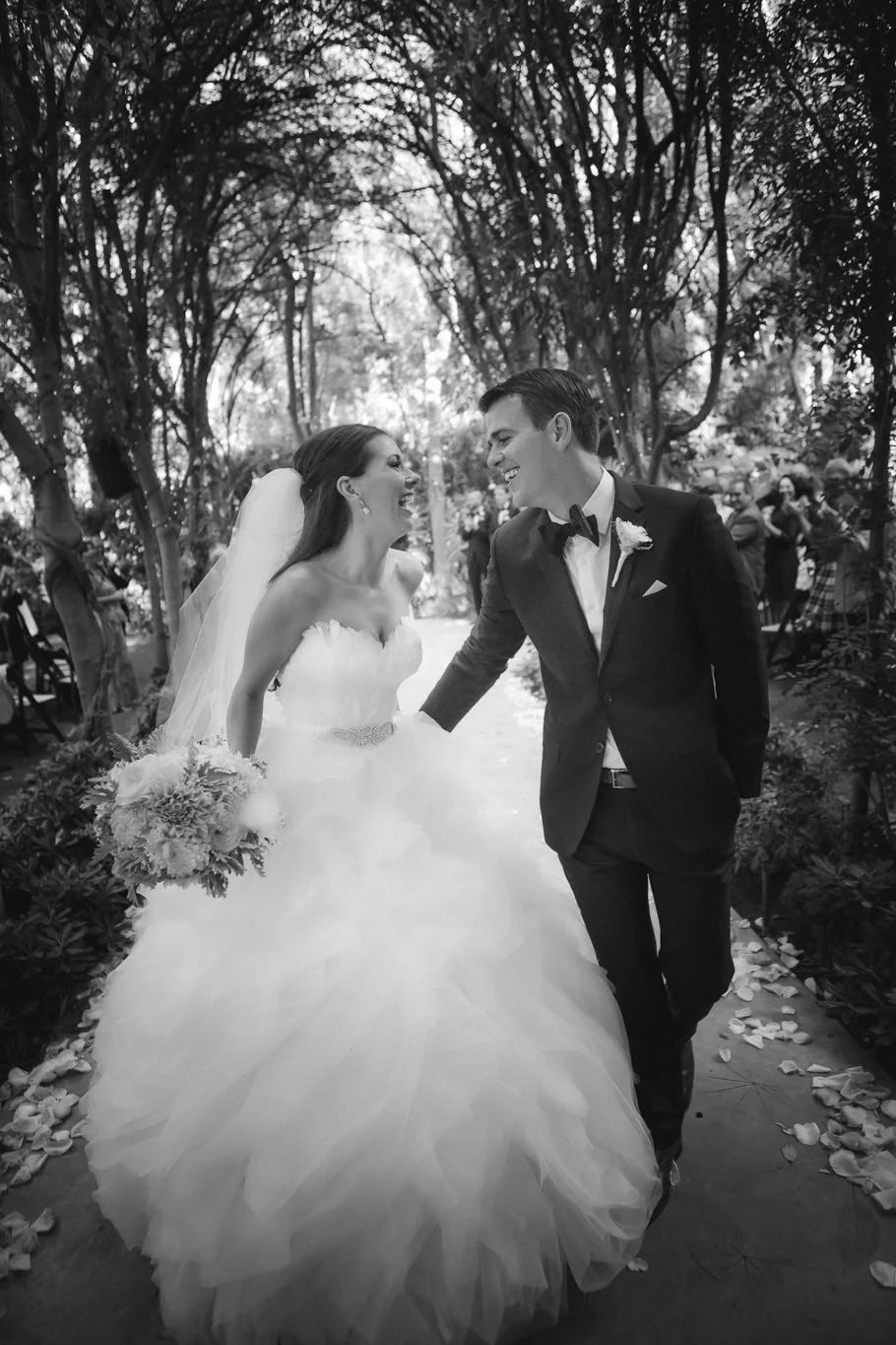 Black and white photo of a bride in a wedding dress and a groom in a suit walking together, smiling and laughing outdoors surrounded by trees and guests.