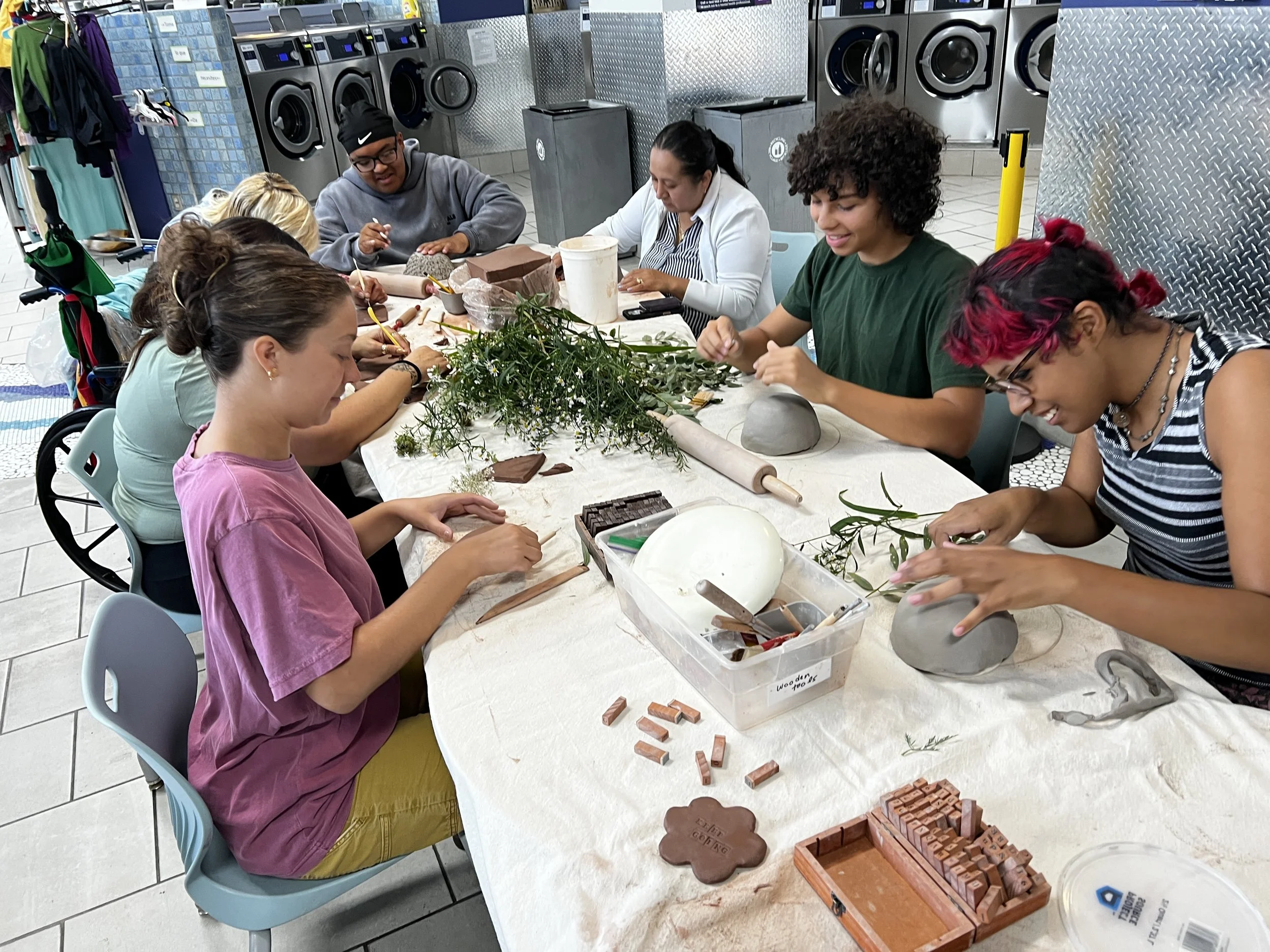 People sitting around a table engaged in crafting with natural materials like branches and leaves, with various crafting tools and materials, including bricks and clay, on the table. In the background, laundry machines are visible in a laundromat.
