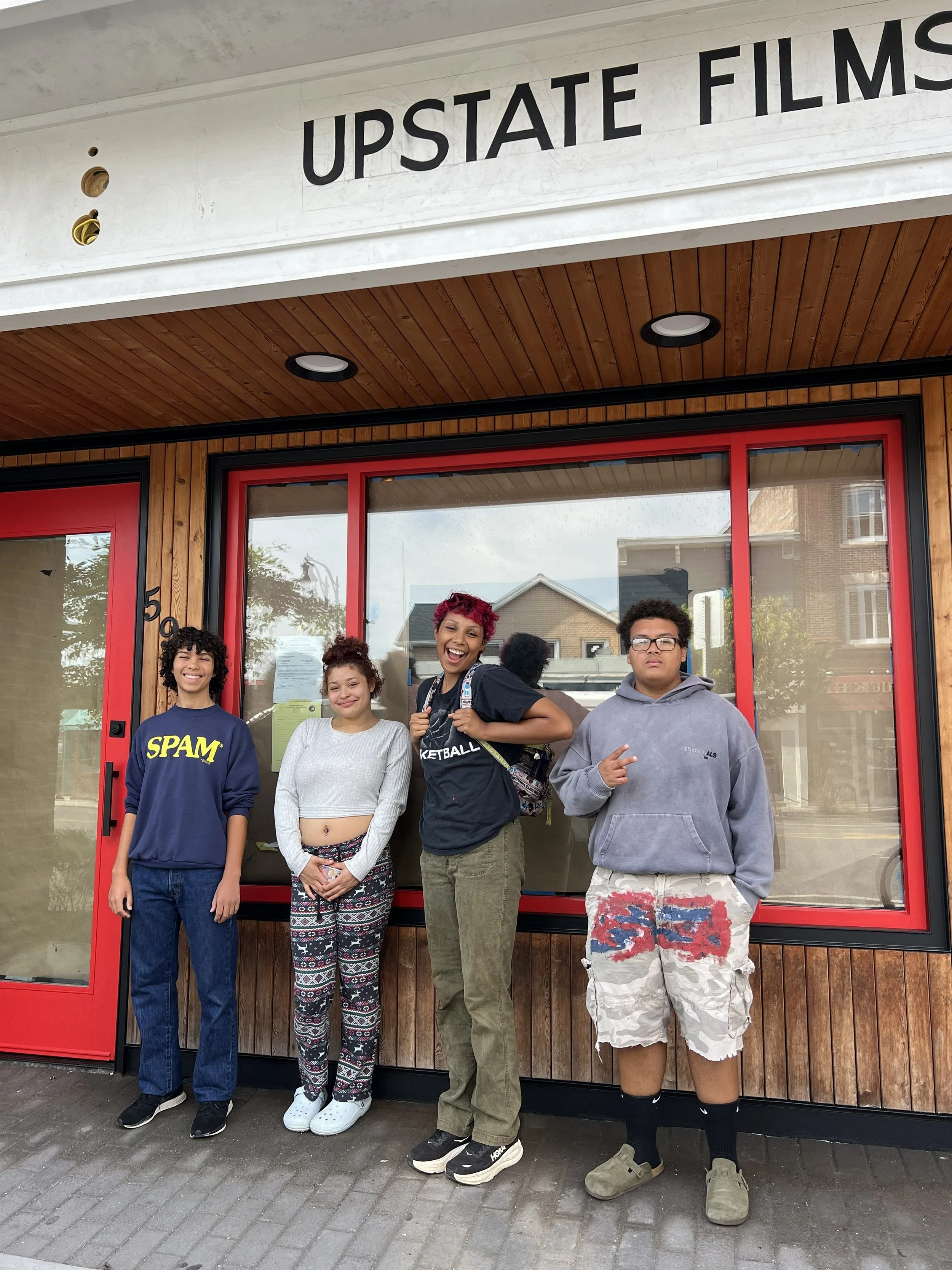 Group of five teenagers standing in front of a storefront with wooden paneling and red door, smiling and posing for a photo.