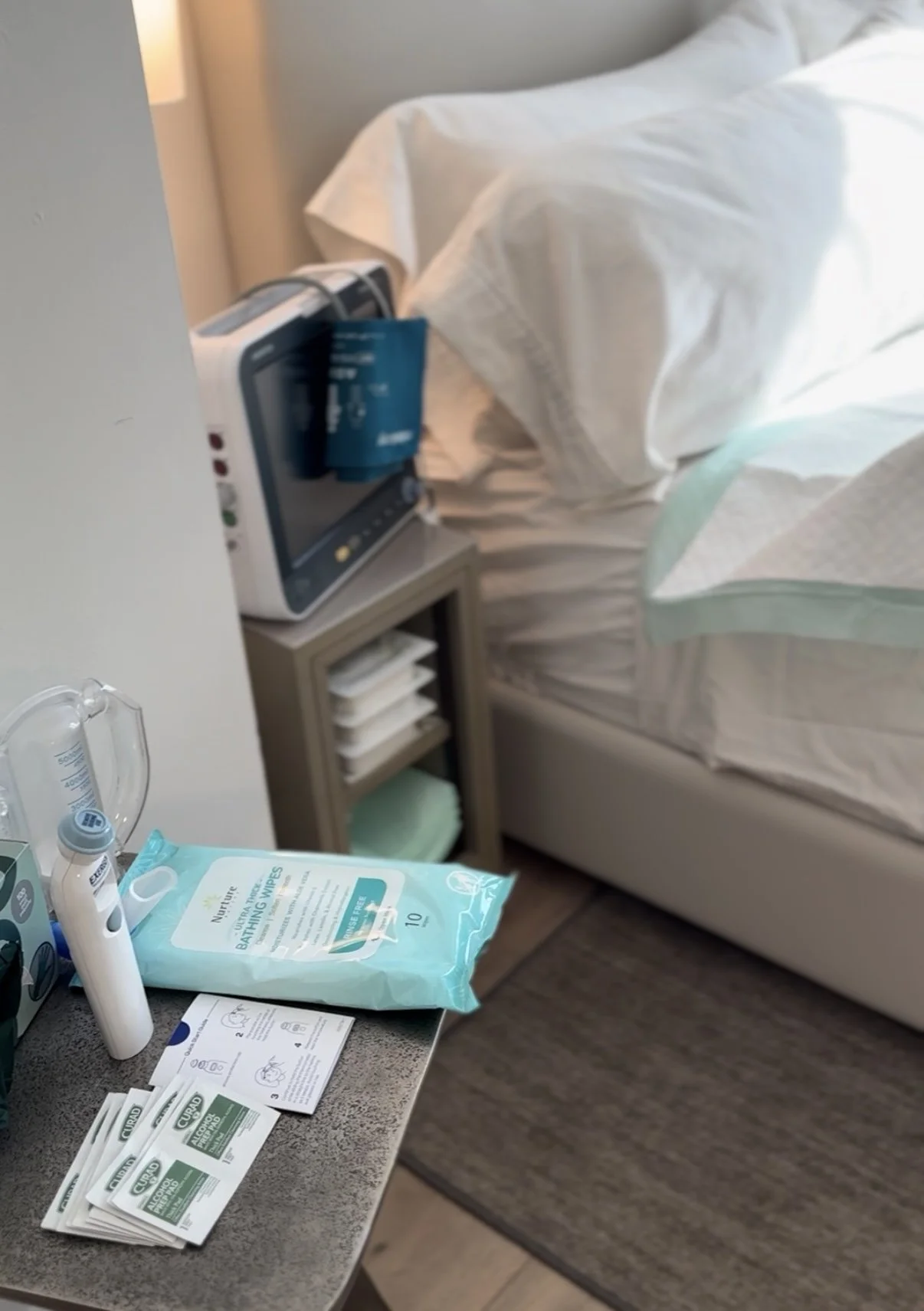 Hospital bedside table with medical supplies, a pack of bathing wipes, a pulse oximeter, and hospital blood pressure cuff and monitor. A hospital bed with white sheets and a bedside monitor are visible in the background.