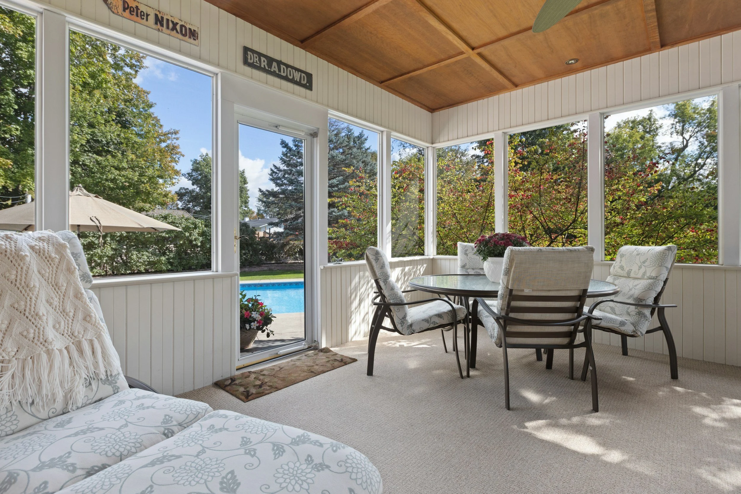 Sunroom with large windows overlooking a backyard pool, with a round dining table and six cushioned chairs, potted plants, and a patterned blanket on a sofa.