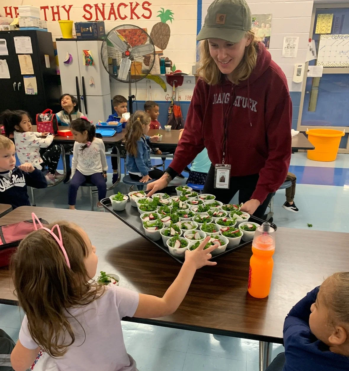 A woman in a red hoodie and green cap distributes small bowls of salad to children in a classroom. Children are seated at tables, some reaching for the bowls, with a large fan and colorful decorations in the background.
