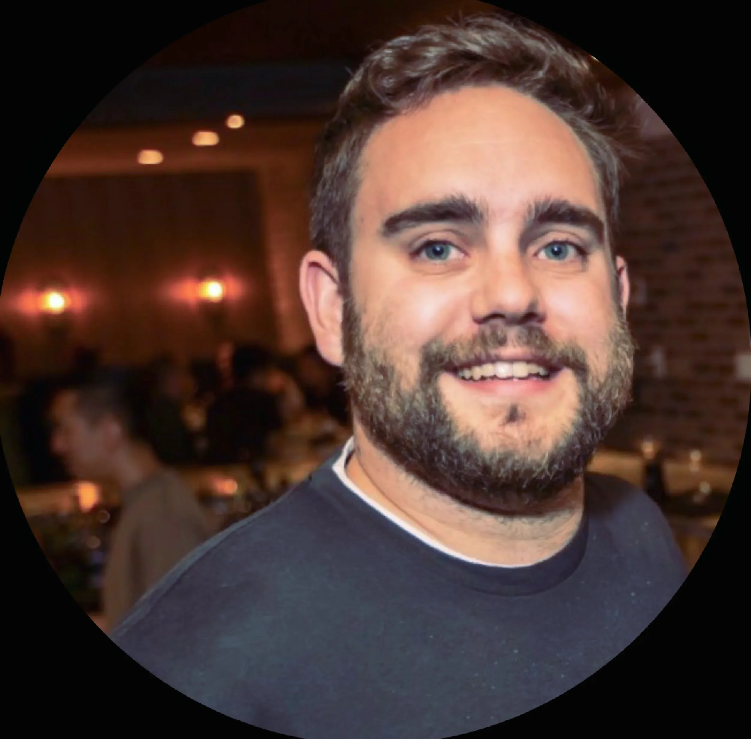 Close-up of a smiling man with blue eyes, beard, and dark hair in a casual setting with warm lighting and a brick wall background.