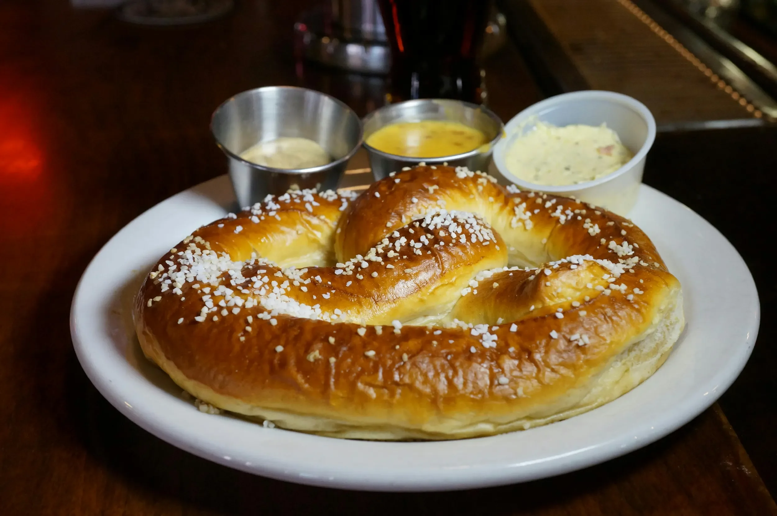 A plate with a large, golden-brown pretzel sprinkled with coarse salt, accompanied by three small containers of dipping sauces or spreads.