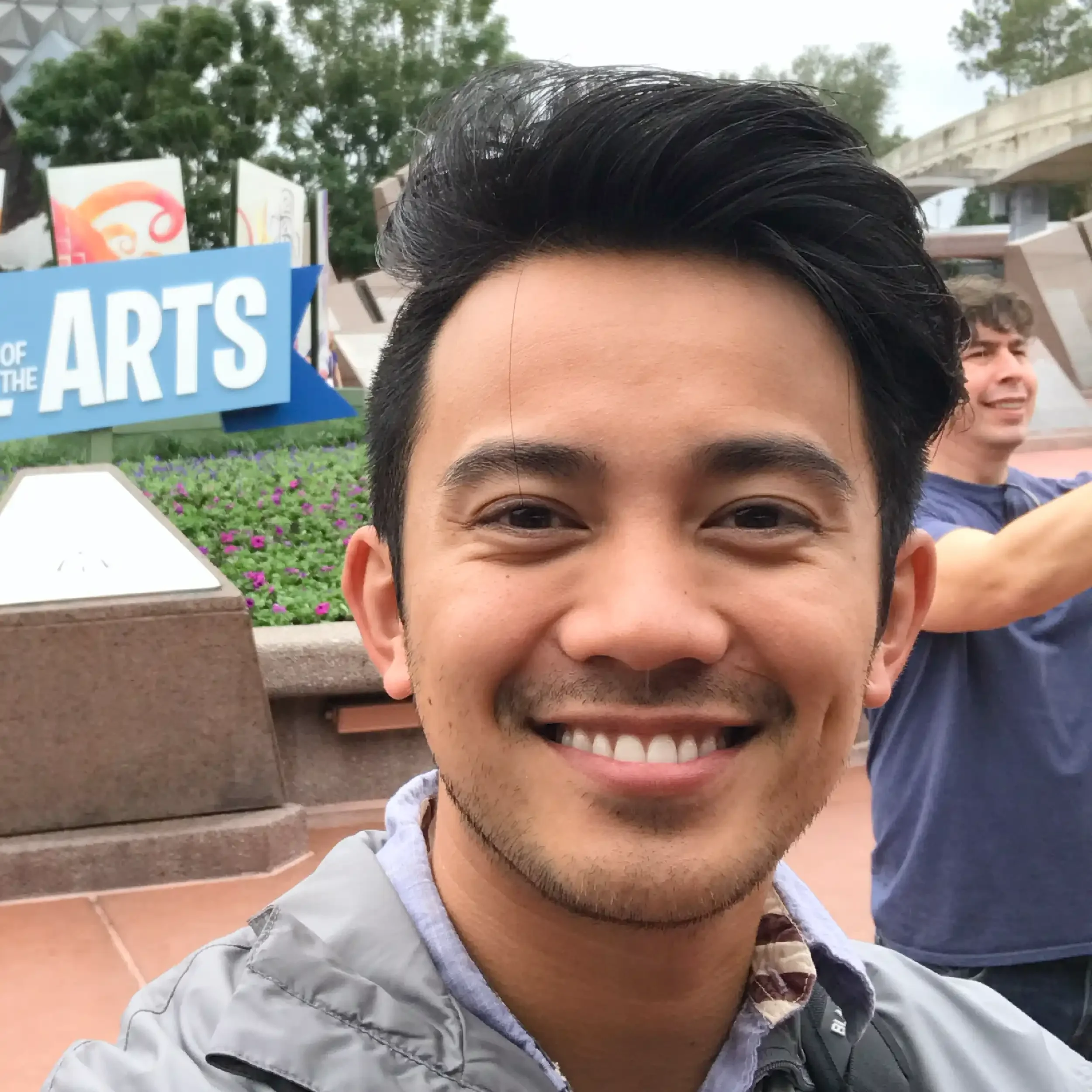 A smiling young man taking a selfie outdoors, with a man in a blue shirt behind him, an arts-themed sign, and purple flowers in the background.