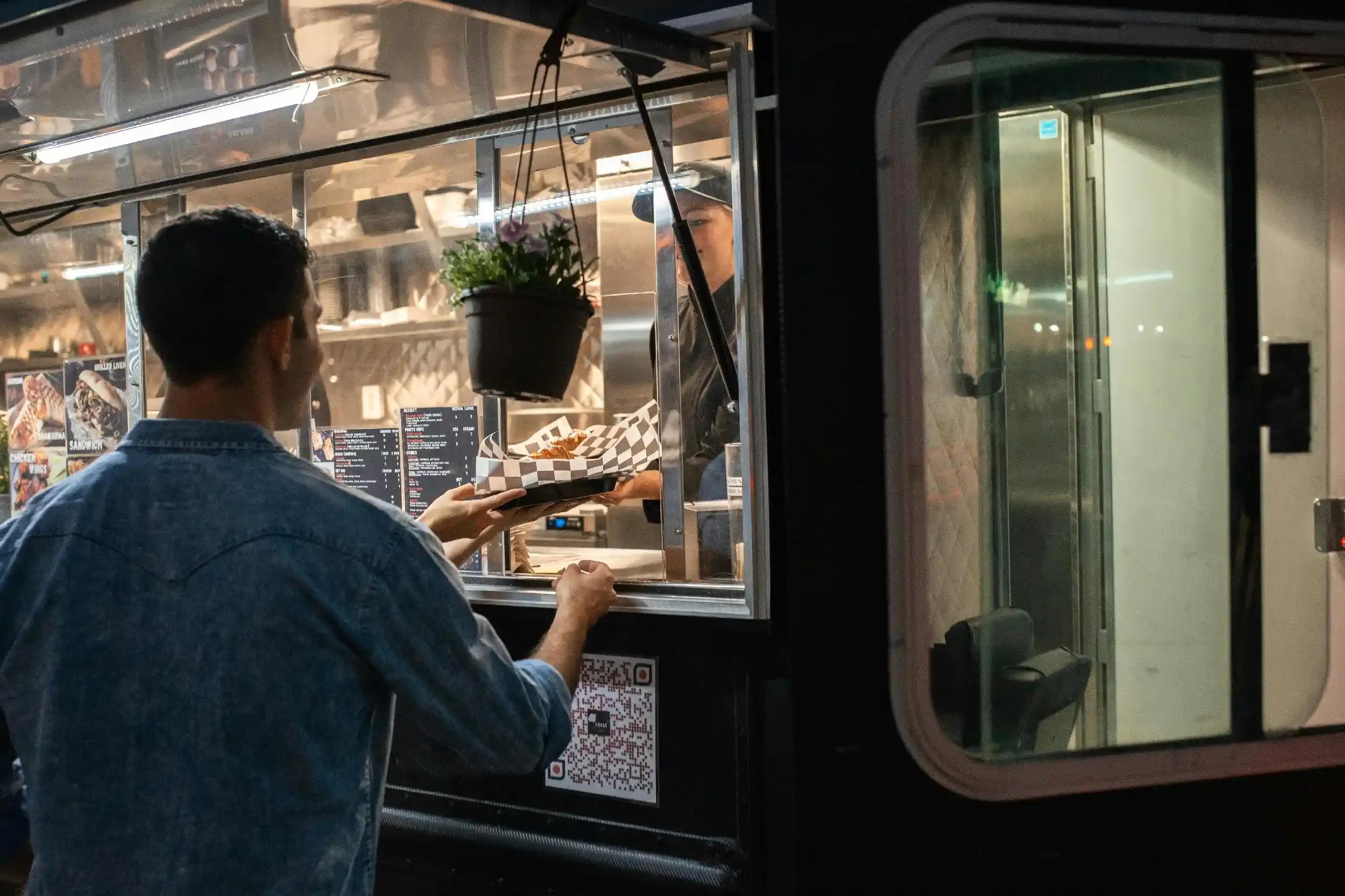 A young man in a denim jacket orders food at a food truck window. The food truck has a menu displayed inside, with a person serving food behind the window. A potted plant hangs outside the window, and a QR code is visible below.