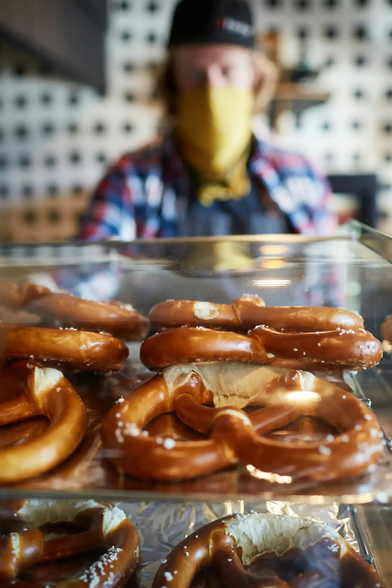 Display of pretzels in a bakery, with a person in a plaid shirt, yellow face mask, and cap in the background.