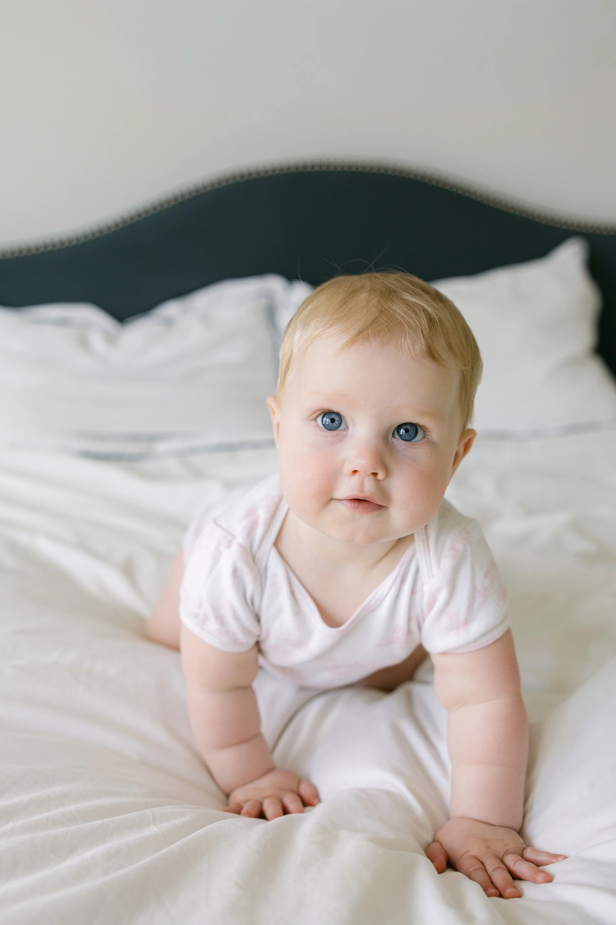 A young child with blonde hair and blue eyes crawling on a bed with white bedding, looking up at the camera.