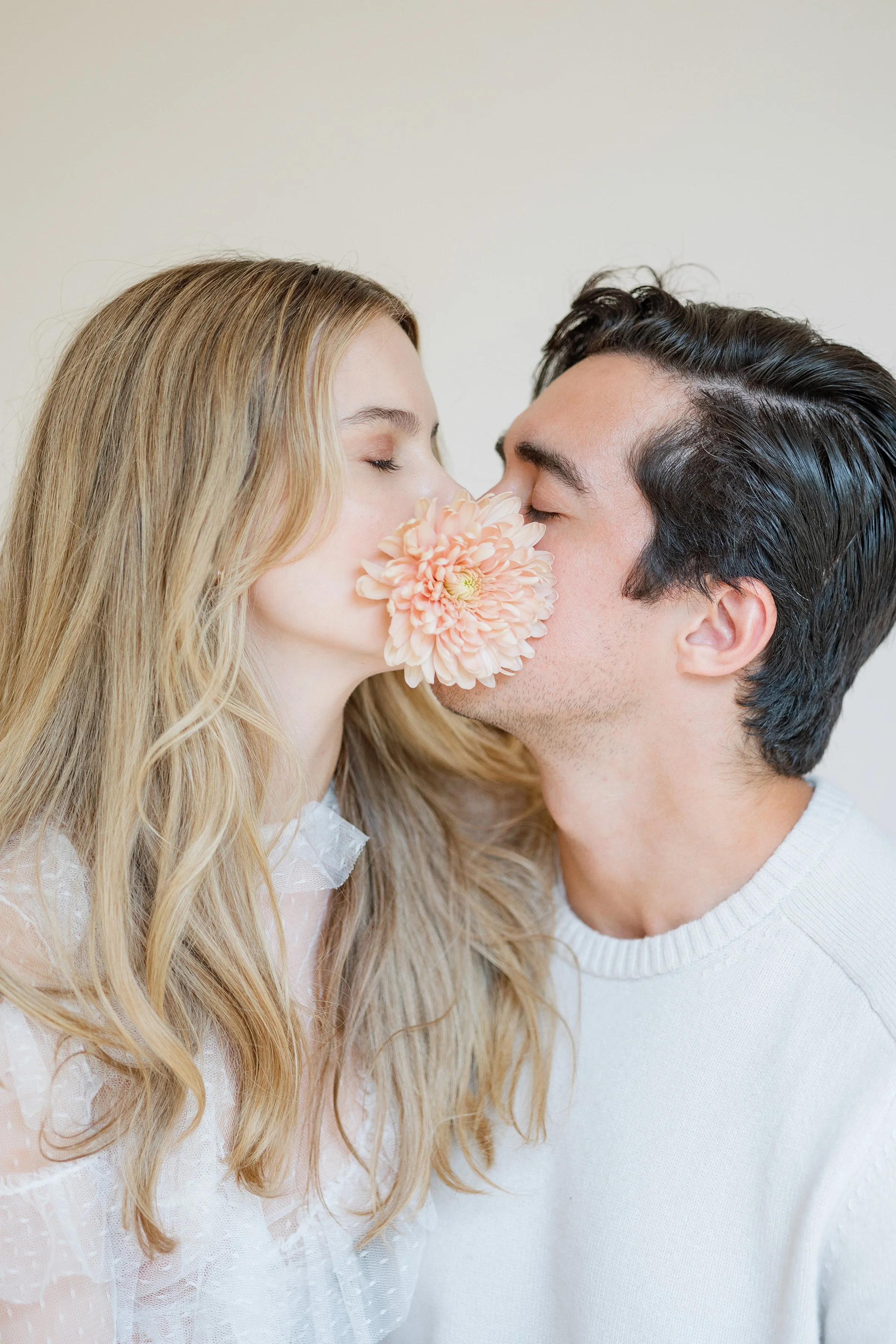 A couple sharing a kiss, with the woman's face partly obscured by a pink flower she holds close to her face. The woman has long, wavy blonde hair and is wearing a white lace dress. The man has dark, wavy hair and is wearing a white sweater.