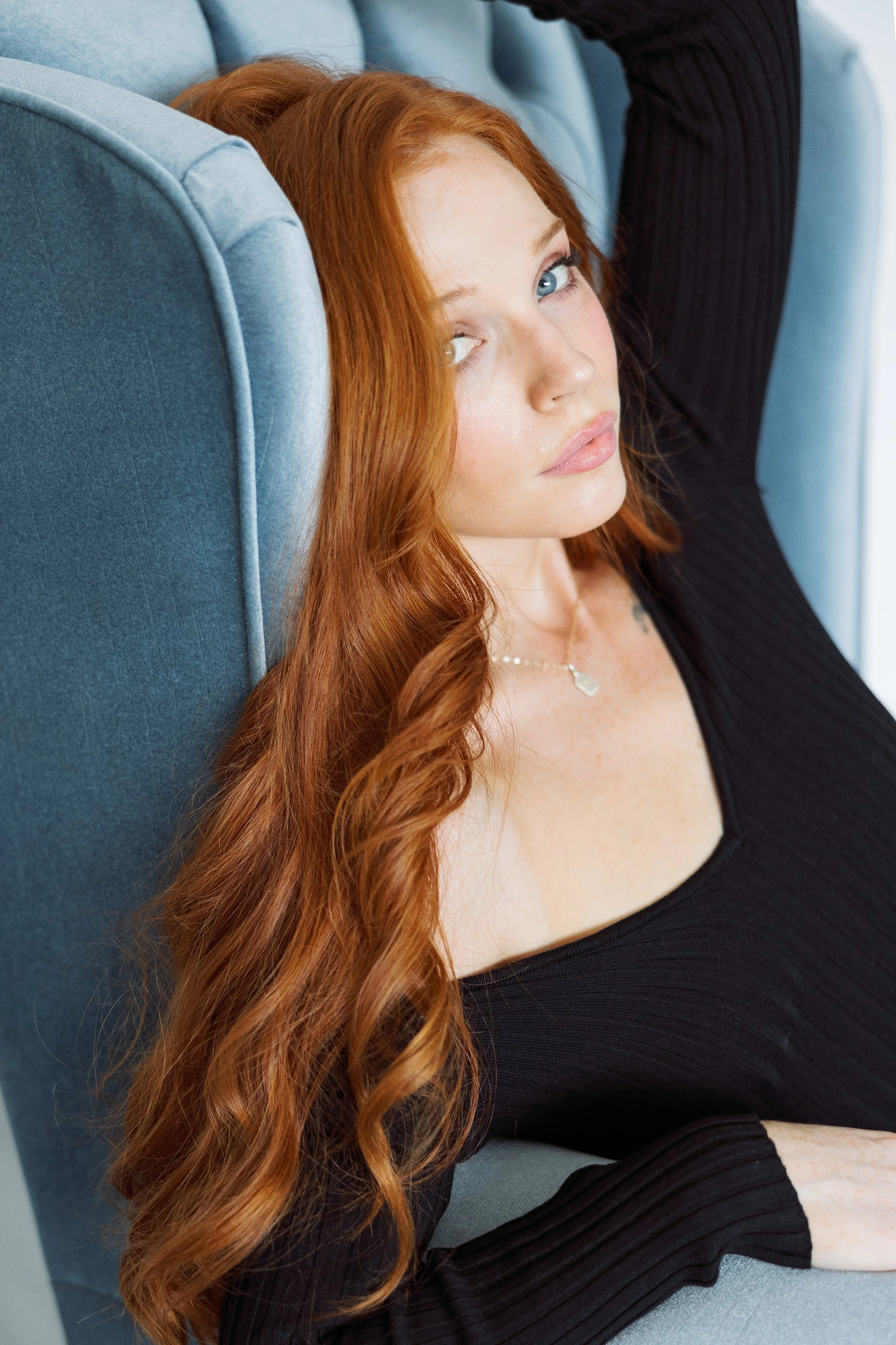 A woman with long red hair, blue eyes, and light skin laying on a blue couch, wearing a black top, with a neutral expression.